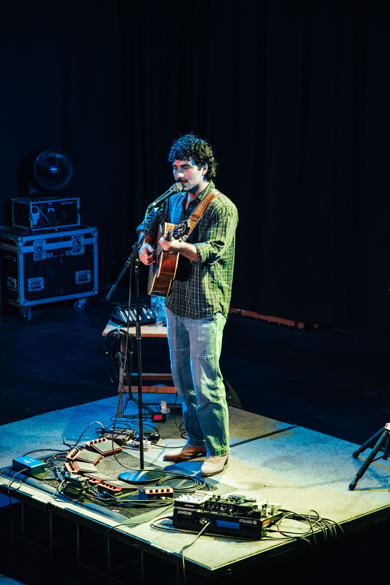 Musician performs on stage with electronic equipment and guitar under dramatic lighting.