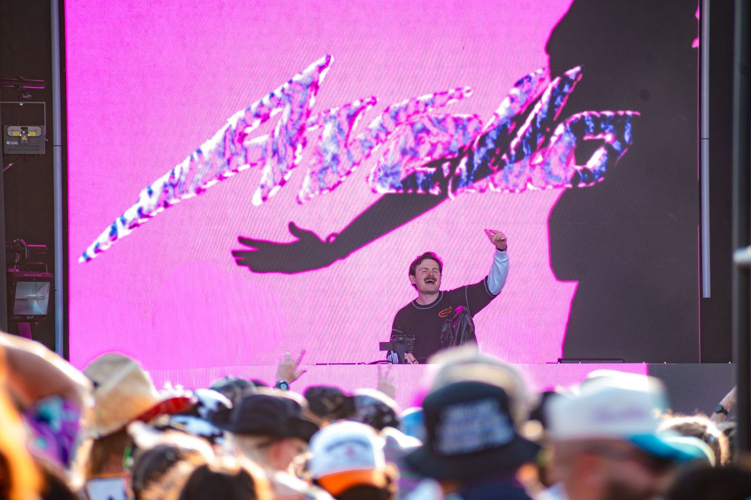 DJ performing on stage at outdoor music festival with pink LED backdrop and large crowd.