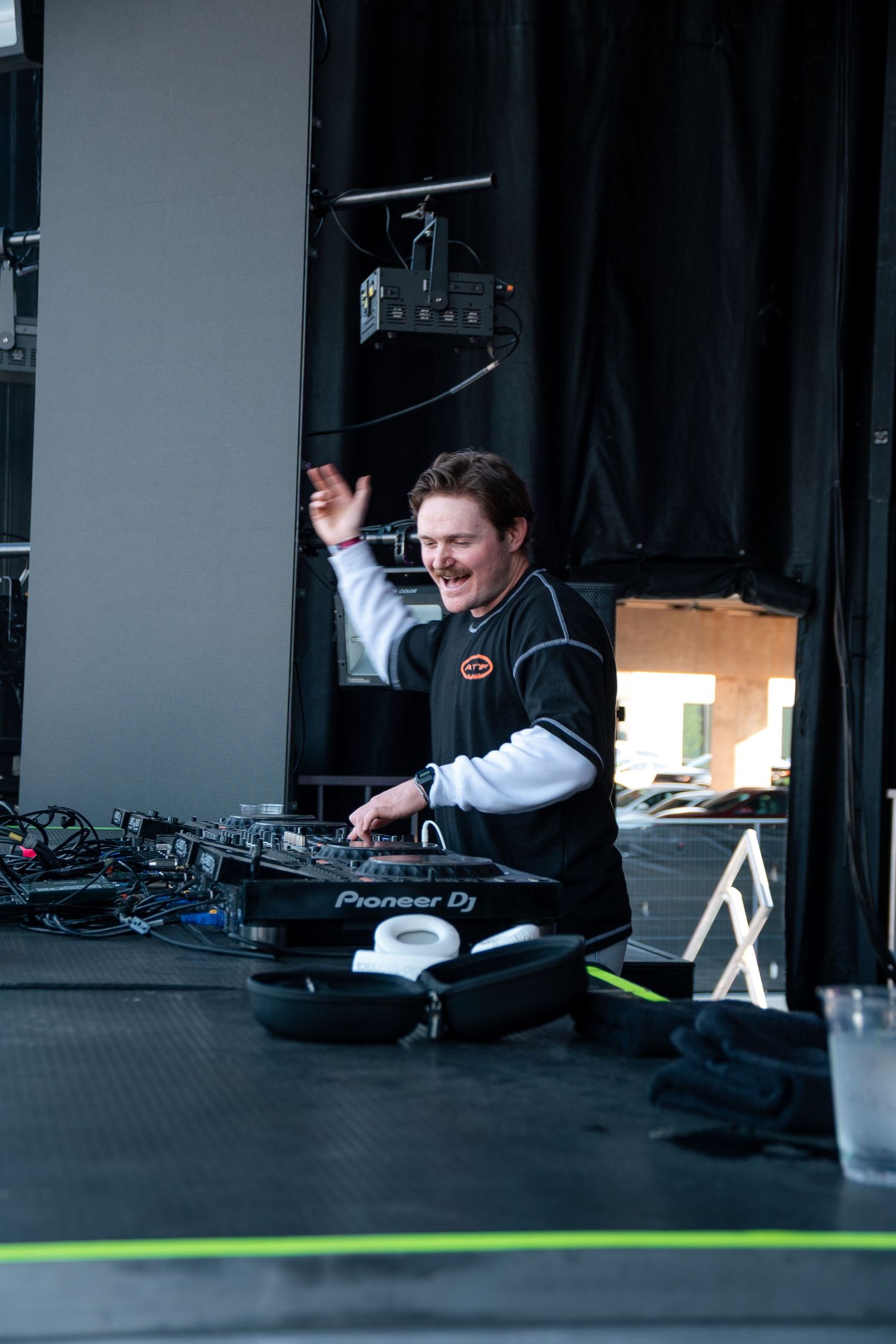 A smiling DJ performs on stage using Pioneer DJ equipment, raising one hand while mixing music outdoors.