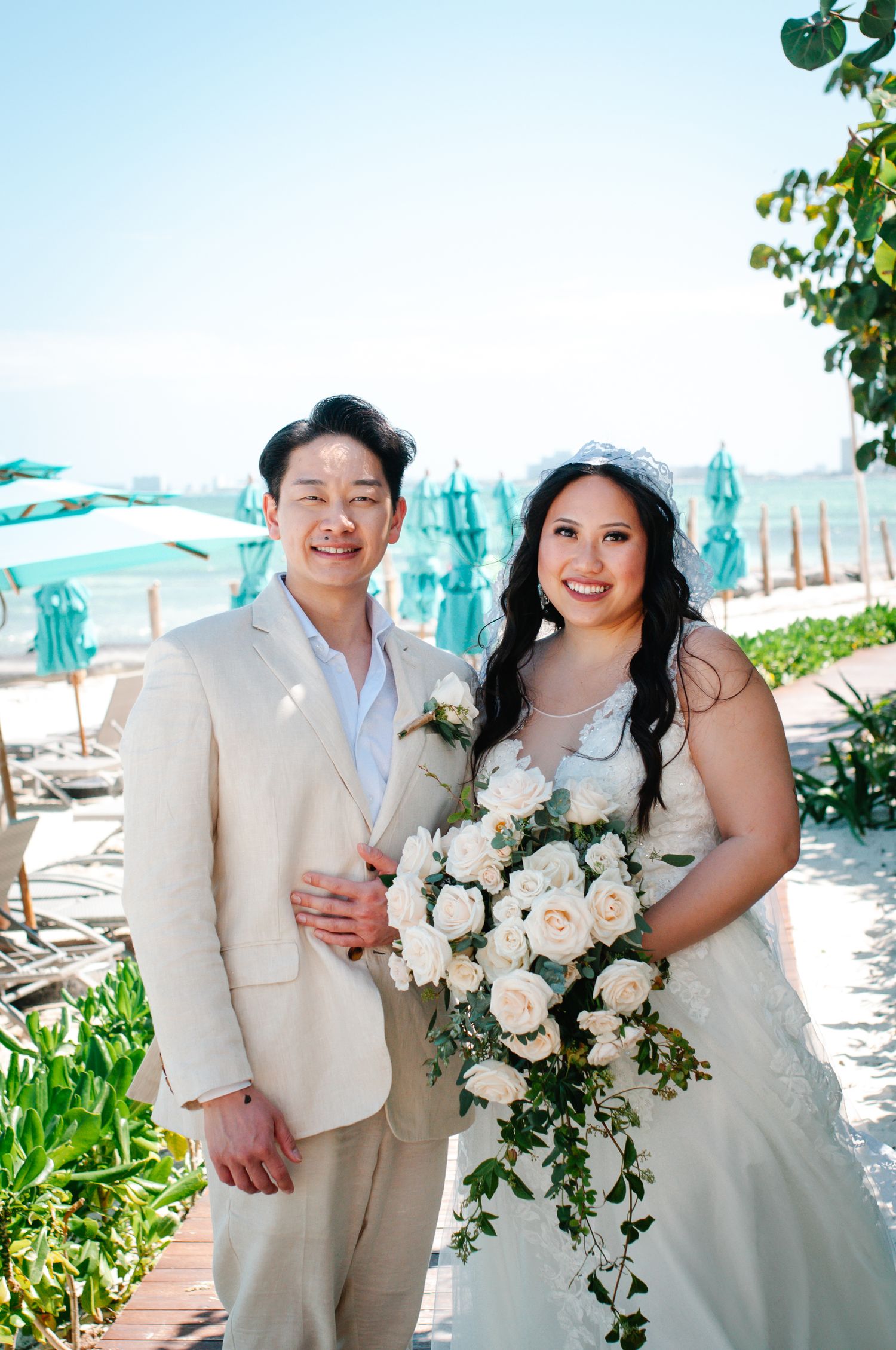 A beach wedding couple poses with a cascading white rose bouquet against turquoise umbrellas and ocean backdrop.