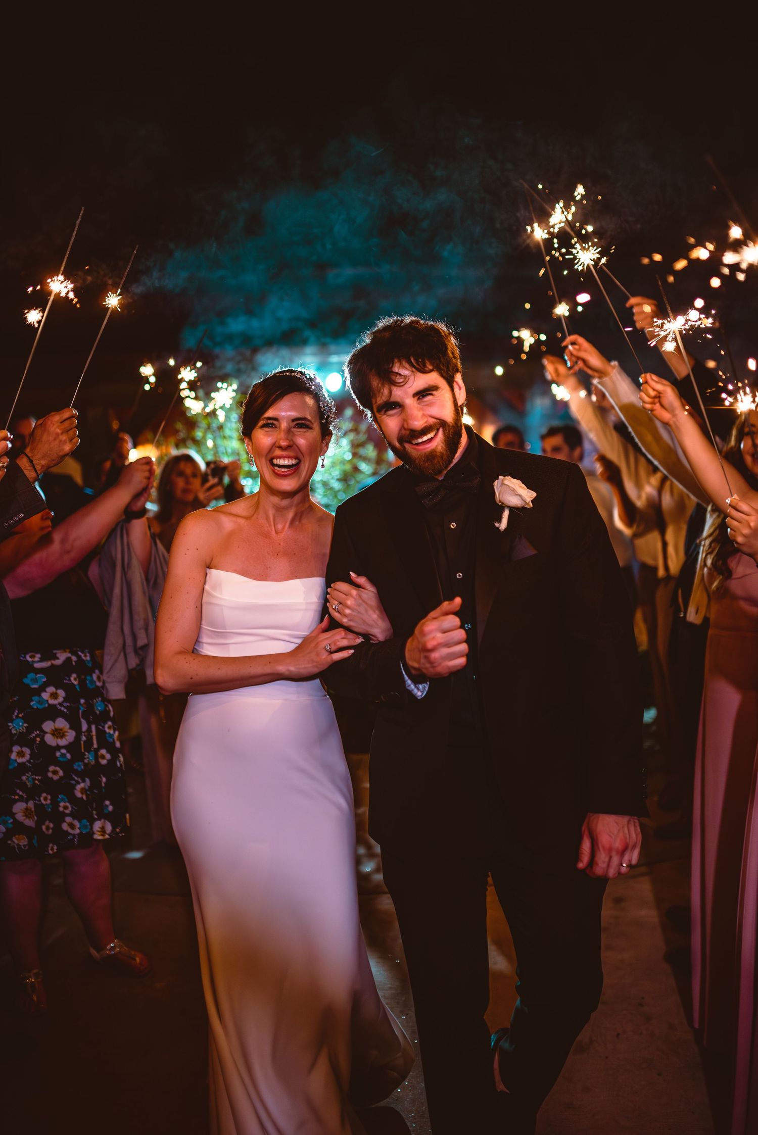 Newlyweds celebrate with sparklers surrounded by wedding guests at night.