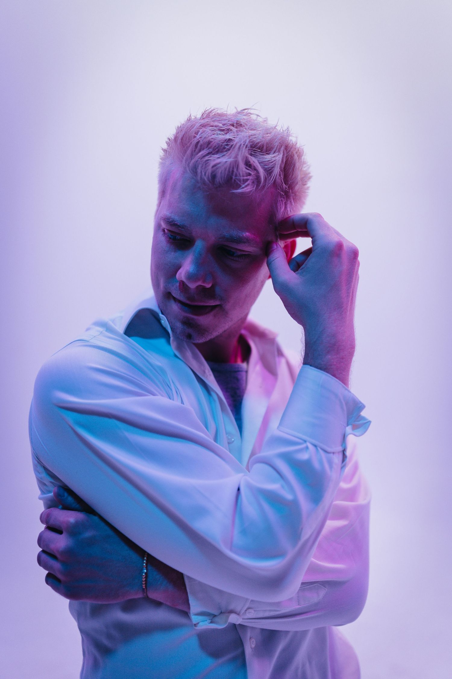 Young man with bleached hair in white shirt poses thoughtfully under purple and pink studio lighting.