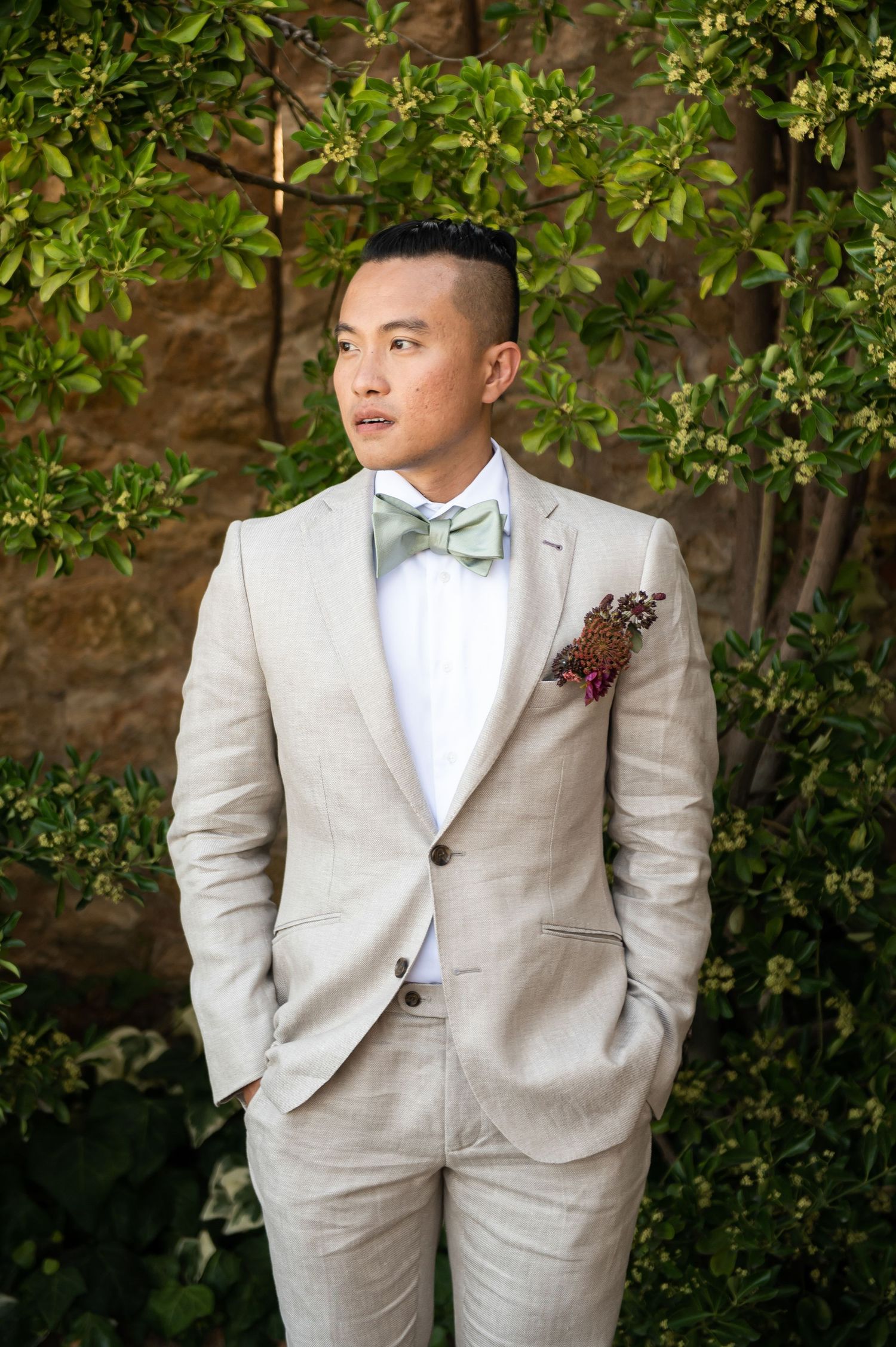 Groom in light beige suit and bow tie poses against greenery backdrop.