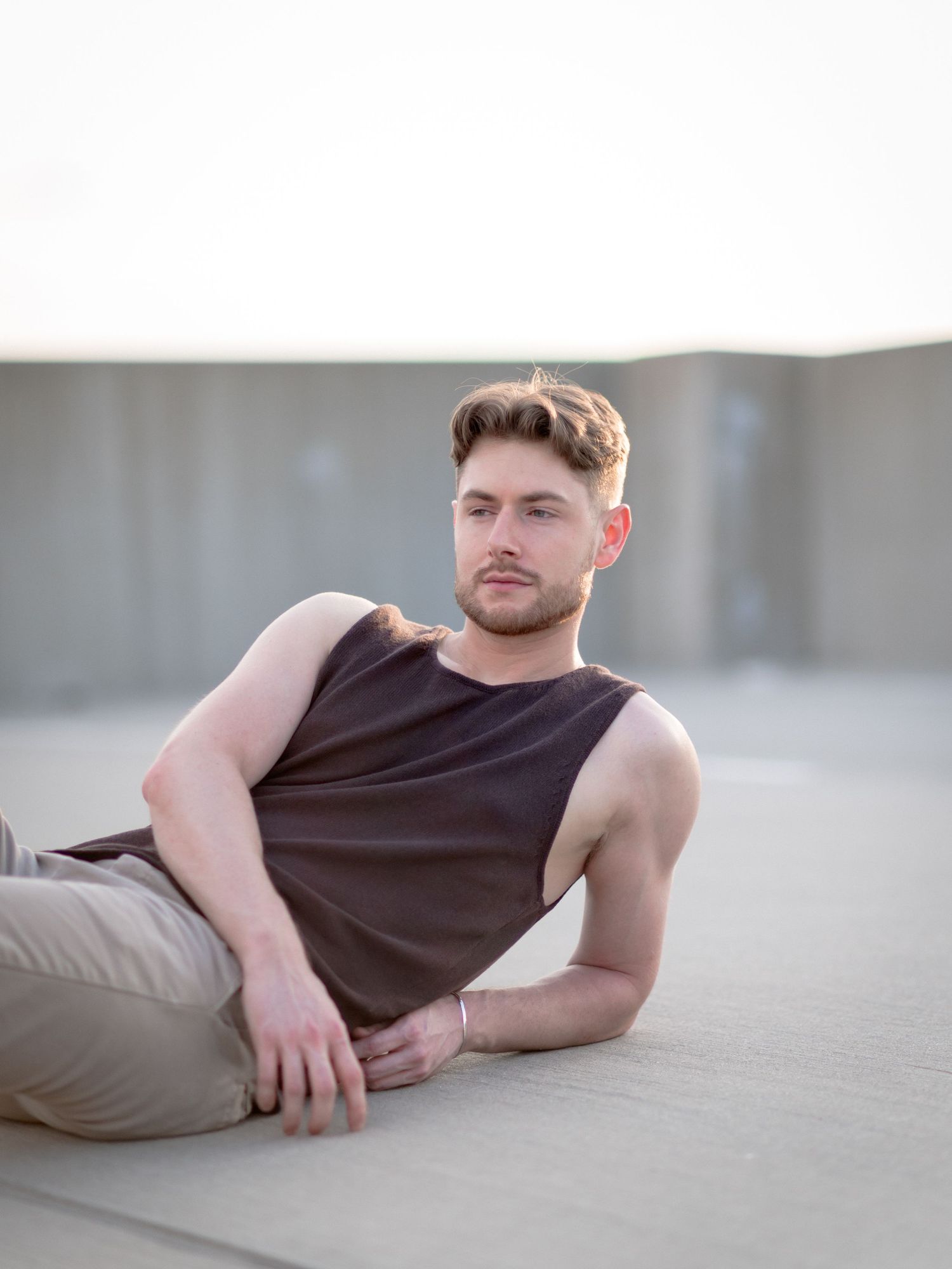 Young man in dark sleeveless shirt and khaki pants relaxing outdoors against a minimalist concrete background.