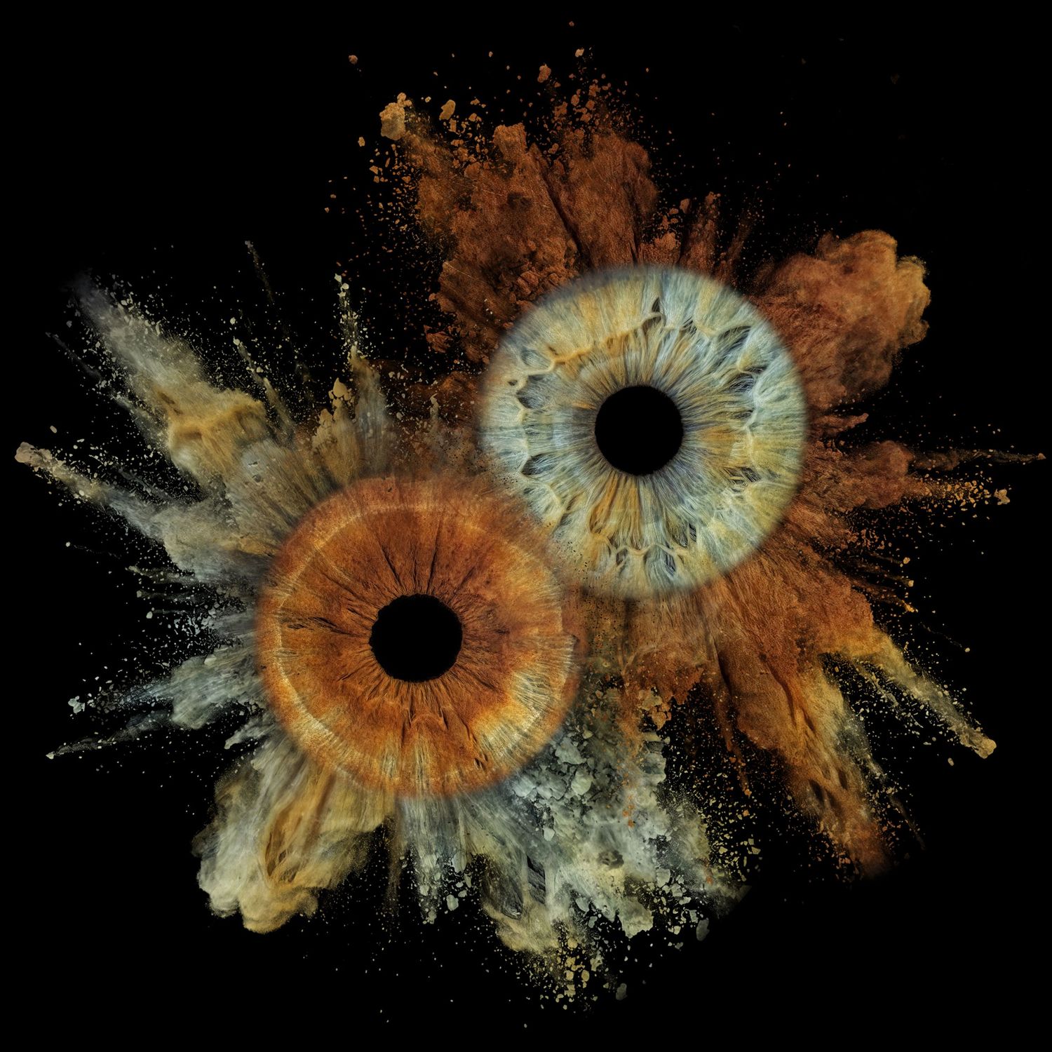 Two human eyes with brown and blue irises surrounded by an explosive burst of golden and amber powder on black background.