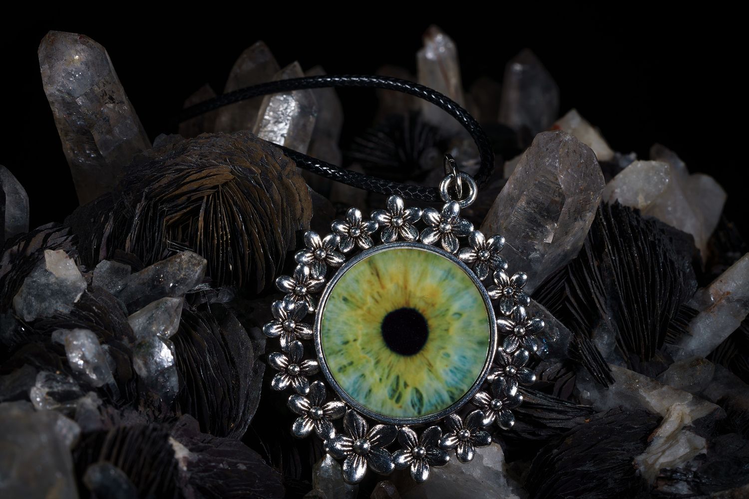 A mysterious green and yellow glass eye pendant with crystal rhinestone surround rests on dark crystals and feathers.