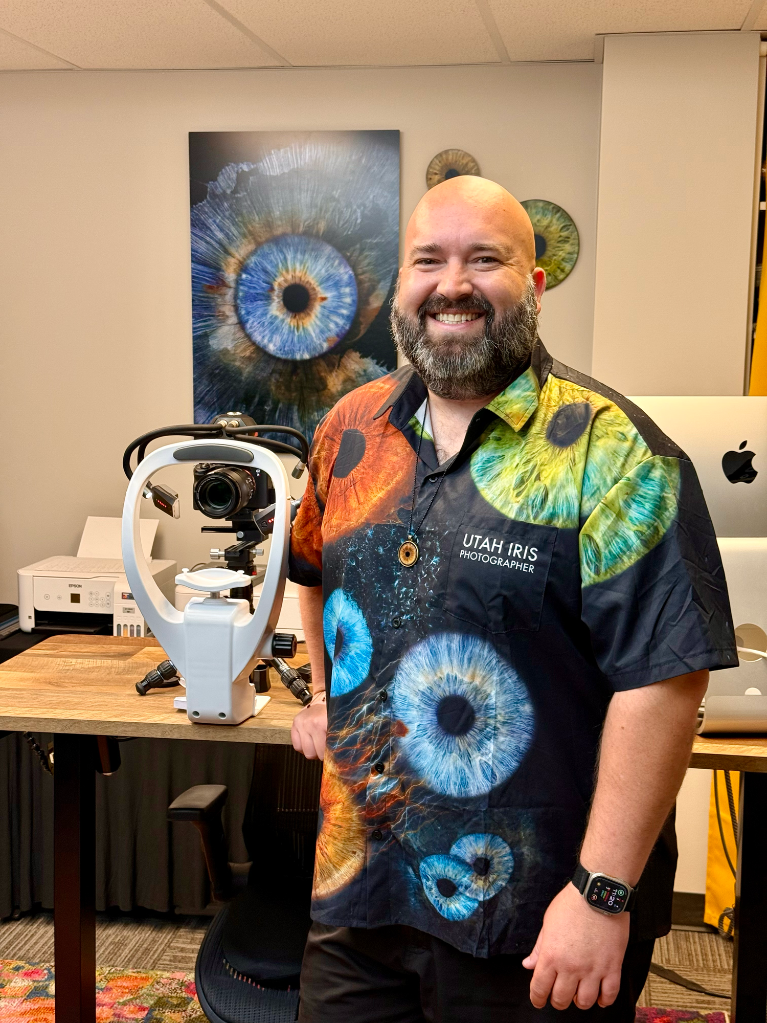 Man in Utah Iris Photography shirt smiles beside eye imaging equipment with colorful iris artwork displayed behind him.