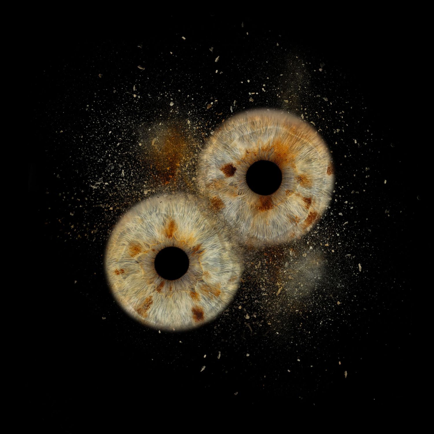 Two human irises exploding against black background, showing golden-brown patterns with scattered particles.