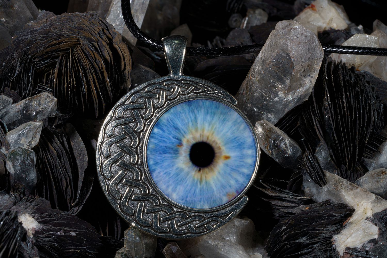 A silver pendant with a realistic glass blue eye centerpiece rests among black feathers and crystal points.