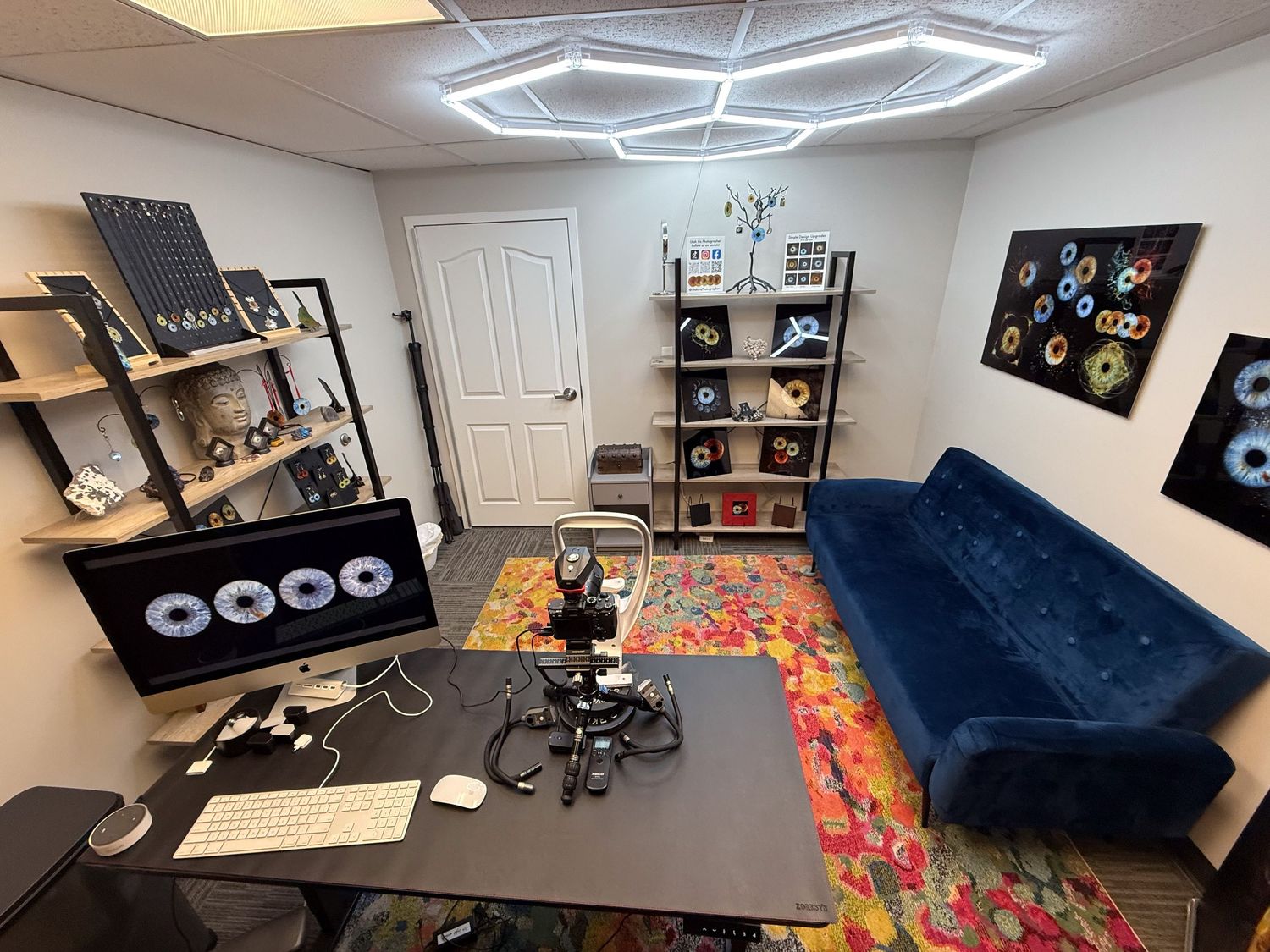 Modern home office with navy blue sofa, geometric ceiling light, shelving units, and colorful area rug under a black desk.