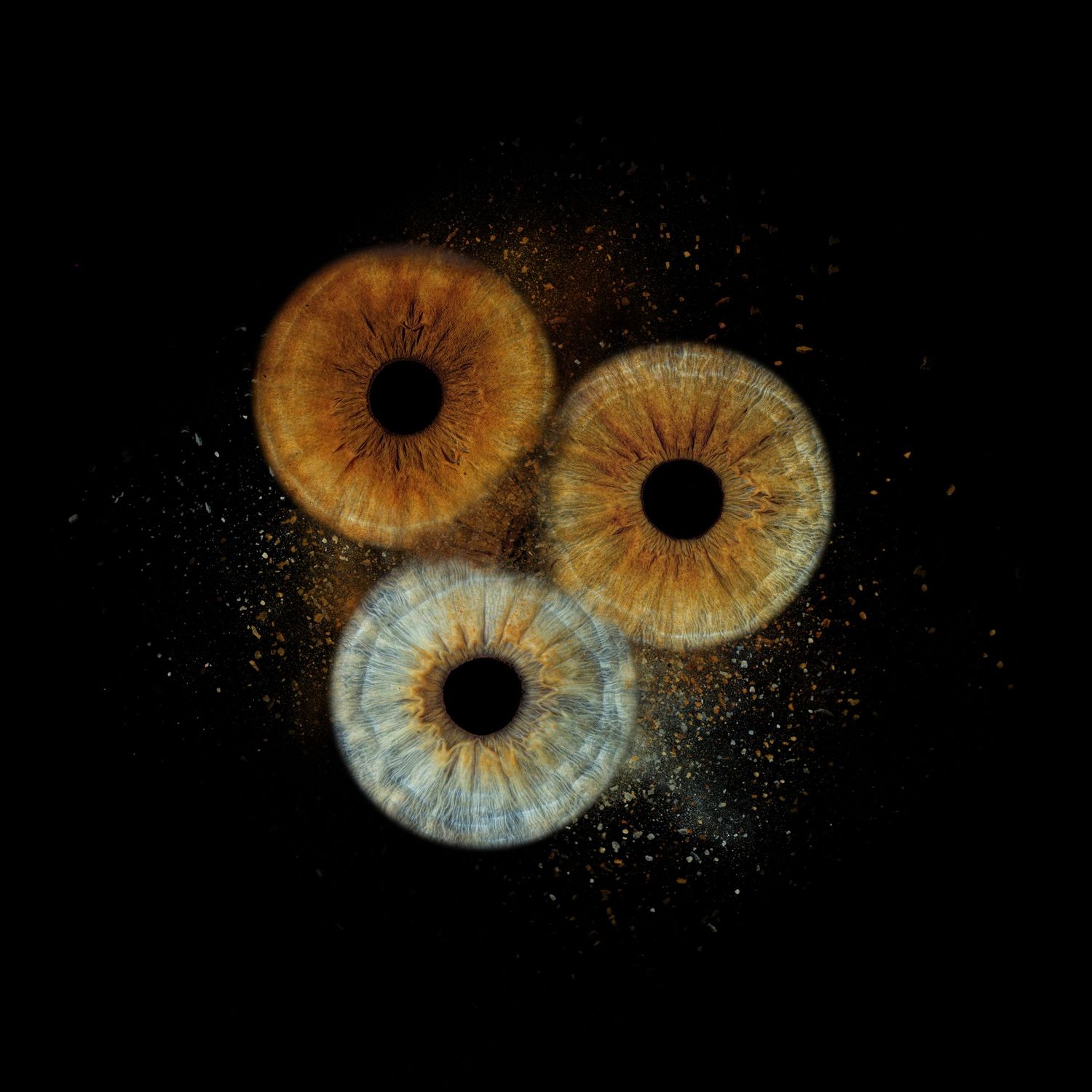 Three human irises in amber, hazel, and blue tones with particle burst effect on black background.