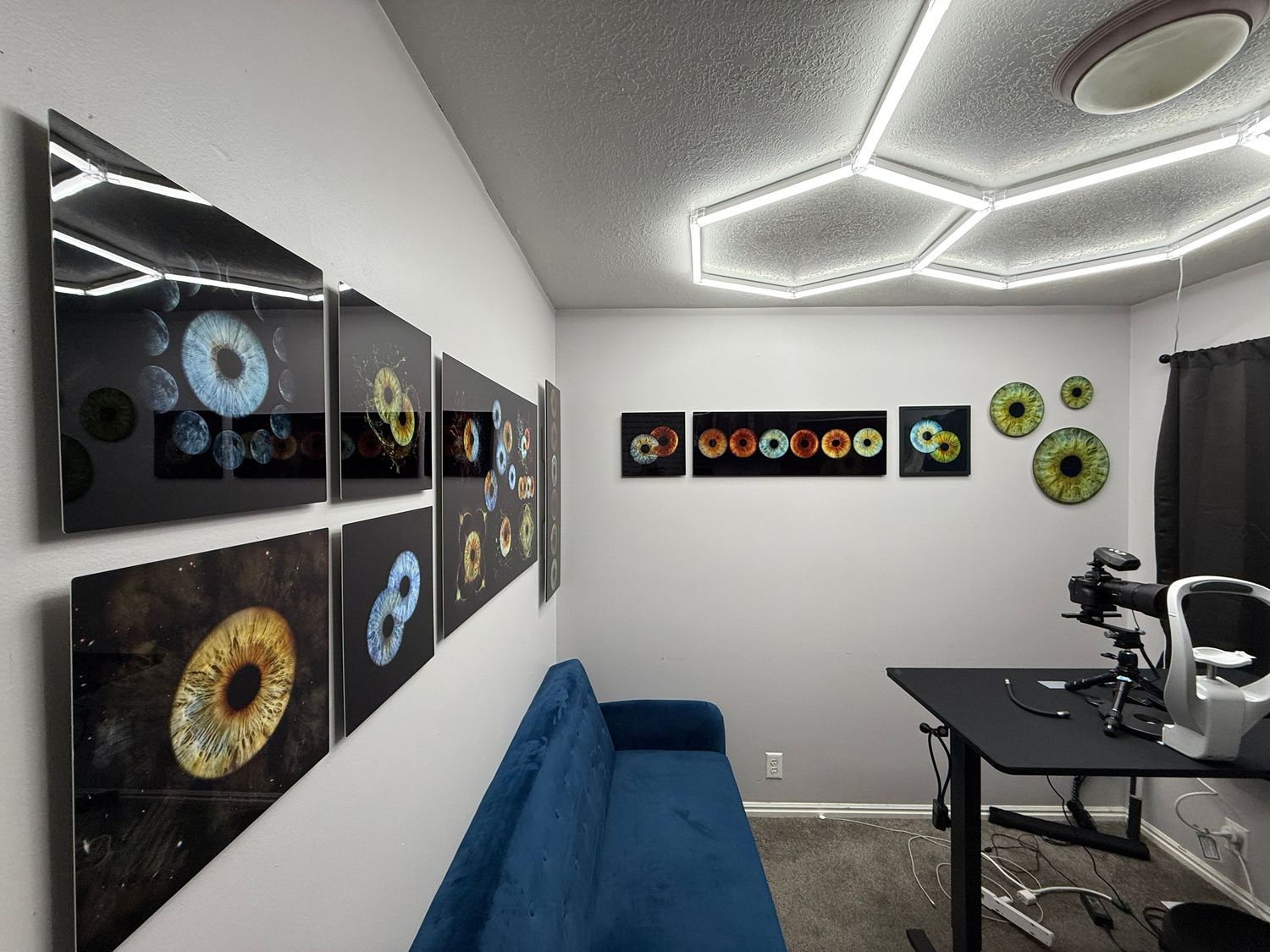 Modern office room with hexagonal LED ceiling lights, colorful eye-themed wall art, blue sofa, and black desk with ergonomic chair.