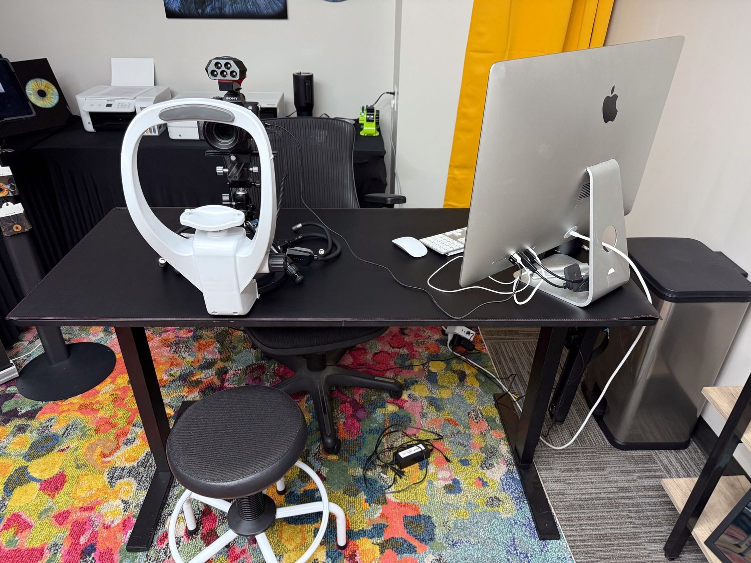 Modern home office setup with iMac computer, gaming headset, and colorful patterned rug on black desk.