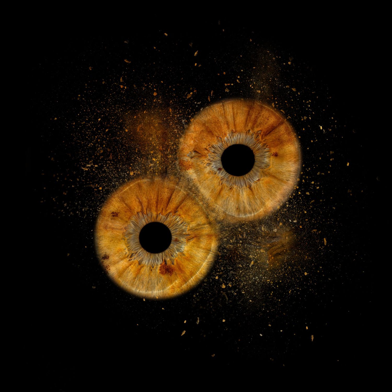 Two golden amber human irises exploding with sparkling particles against a black background.