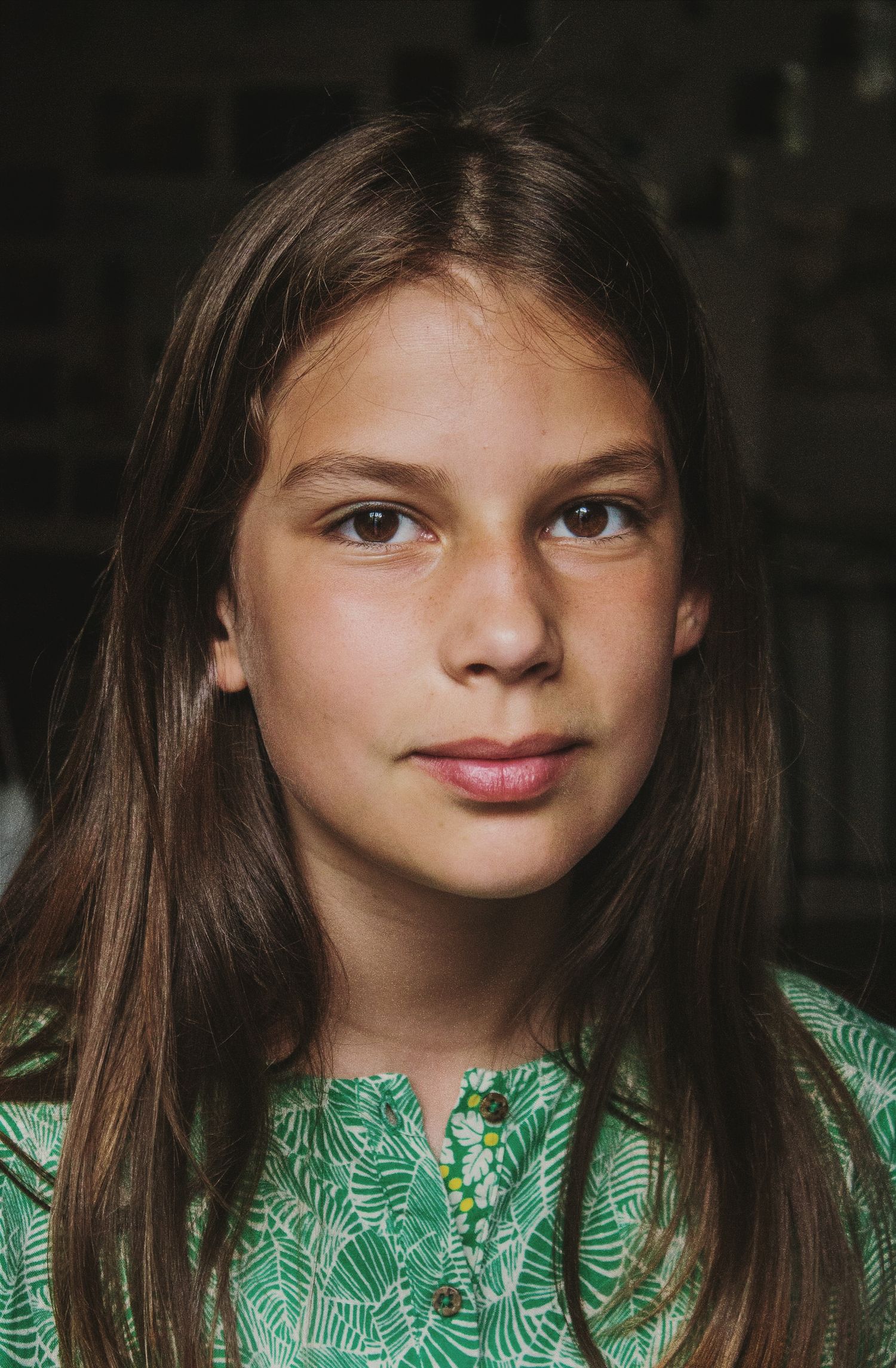 A young girl with long brown hair wearing a green patterned top looks directly at the camera.
