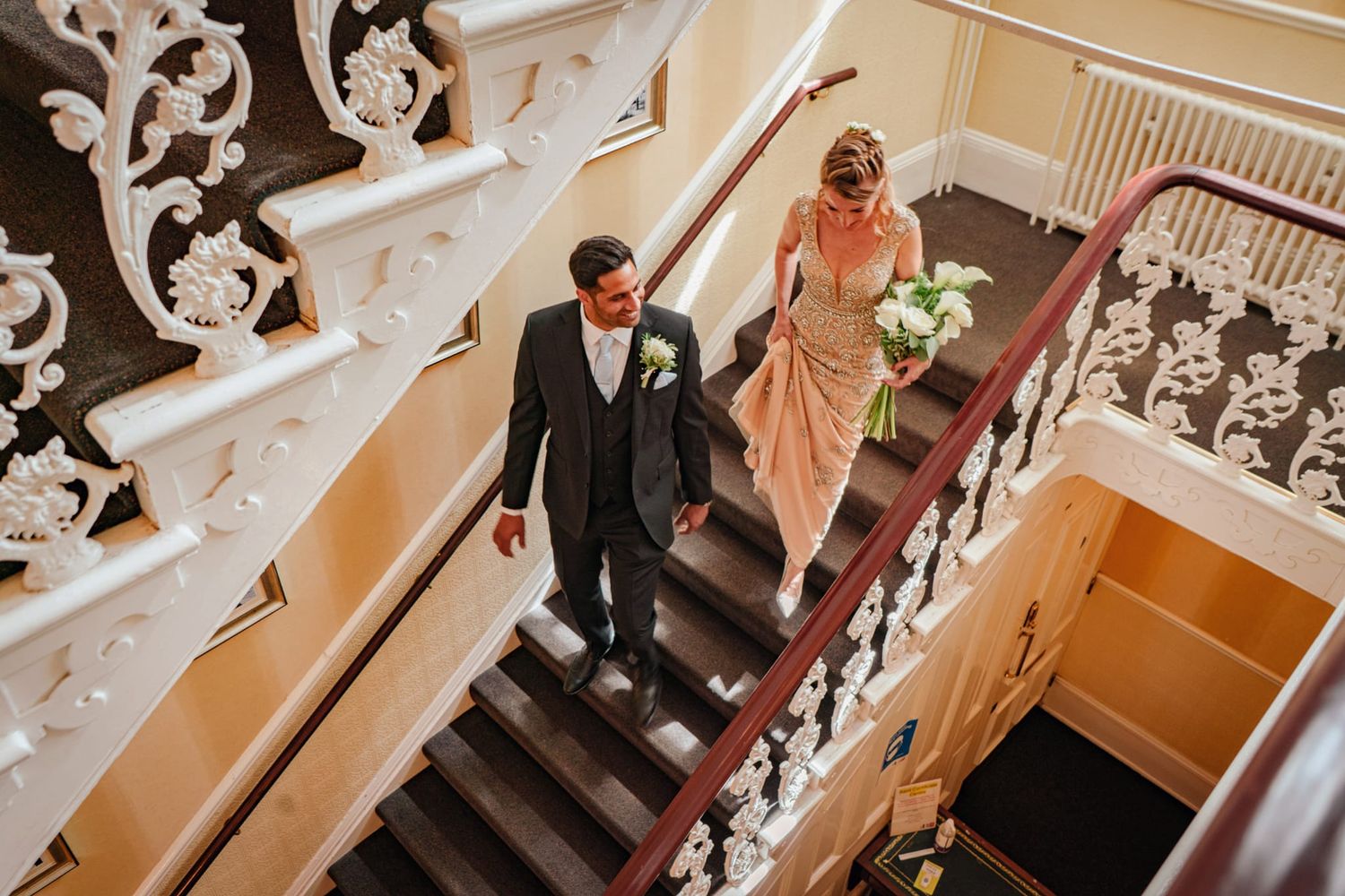 Aerial view of bride and groom walking down an ornate white staircase with decorative railings.