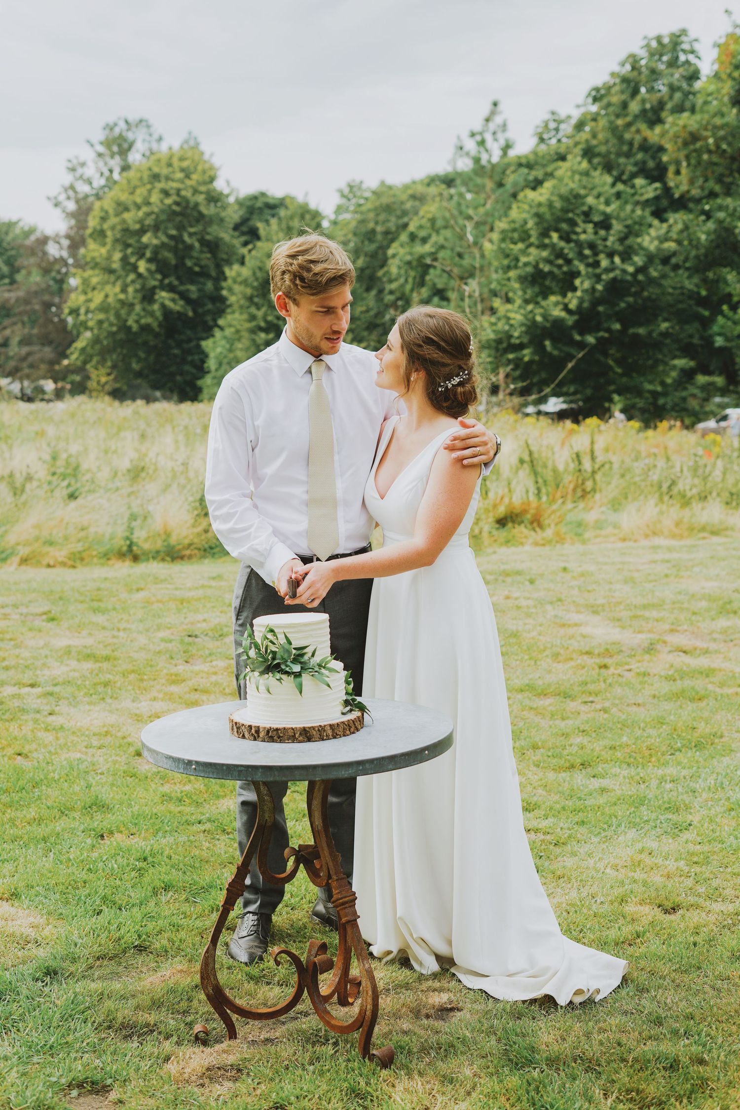 Bride and groom cutting their simple white wedding cake outdoors on a rustic table surrounded by lush greenery.
