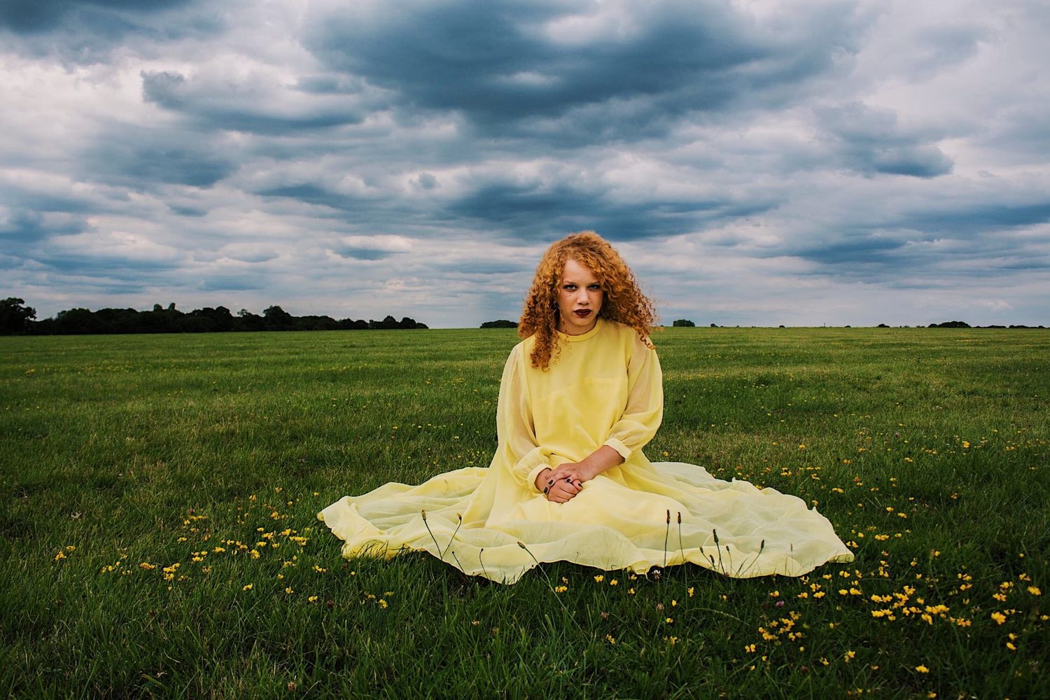 A curly-haired woman in a yellow dress sits cross-legged in a green flower-filled field.