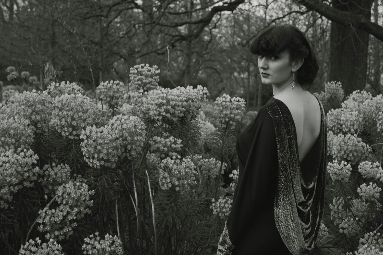 Woman in backless black gown stands among blooming wildflowers in a moody black and white forest setting.