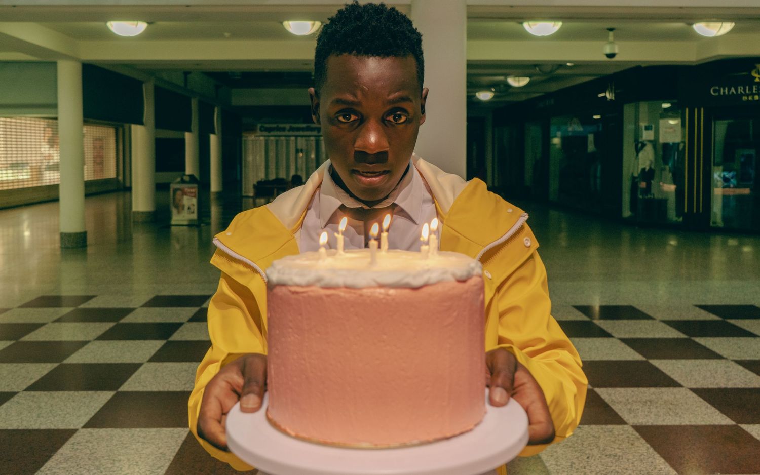Man in yellow jacket holding a pink birthday cake with lit candles in a shopping mall.