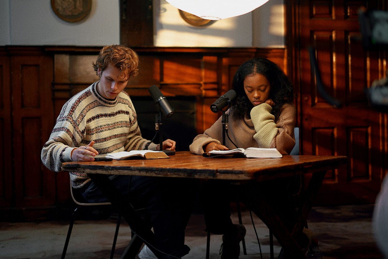 A young man and woman study together at a wooden table in a warmly lit room.