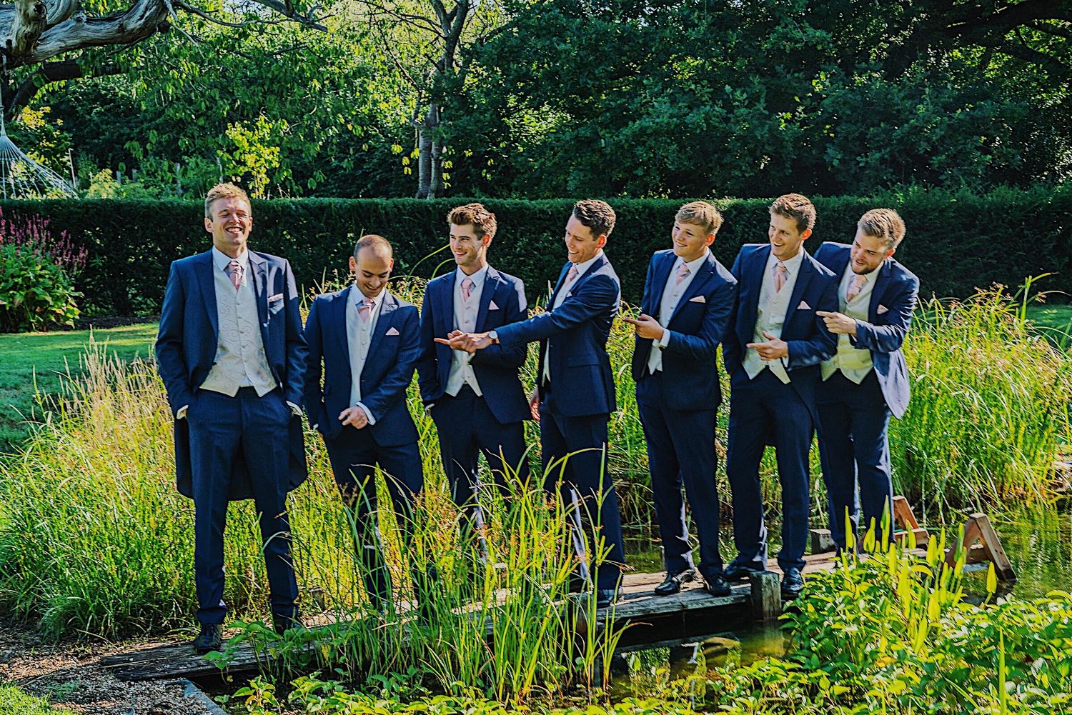 Groom and groomsmen in navy suits posing together by pond in lush garden setting.