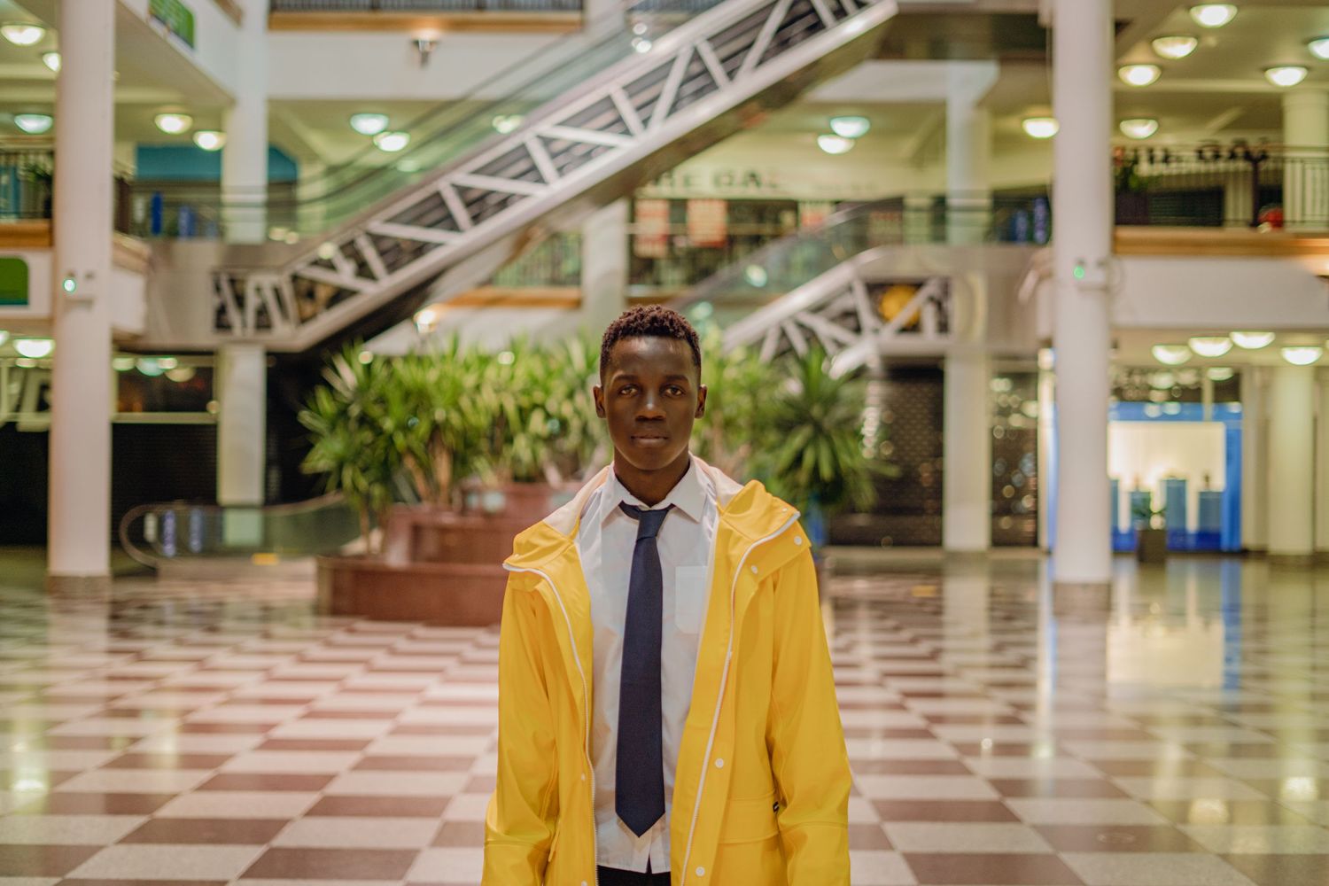 Young man in yellow raincoat and tie standing in a bright modern shopping mall.