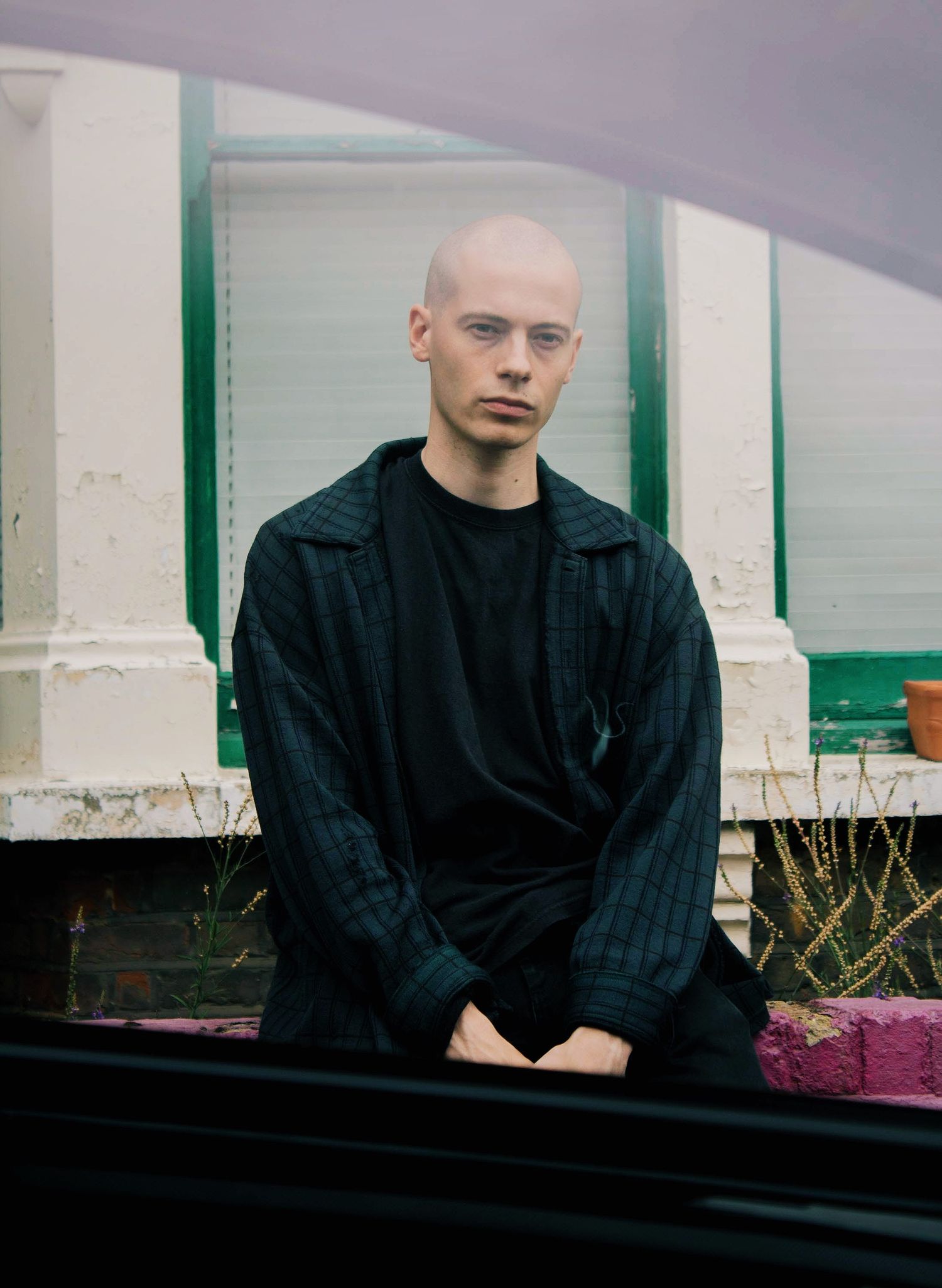 Bald young man in dark plaid jacket sitting in front of a building with green trim.