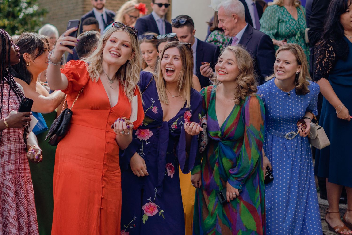Group of joyful female wedding guests in colorful dresses laughing and celebrating outdoors.