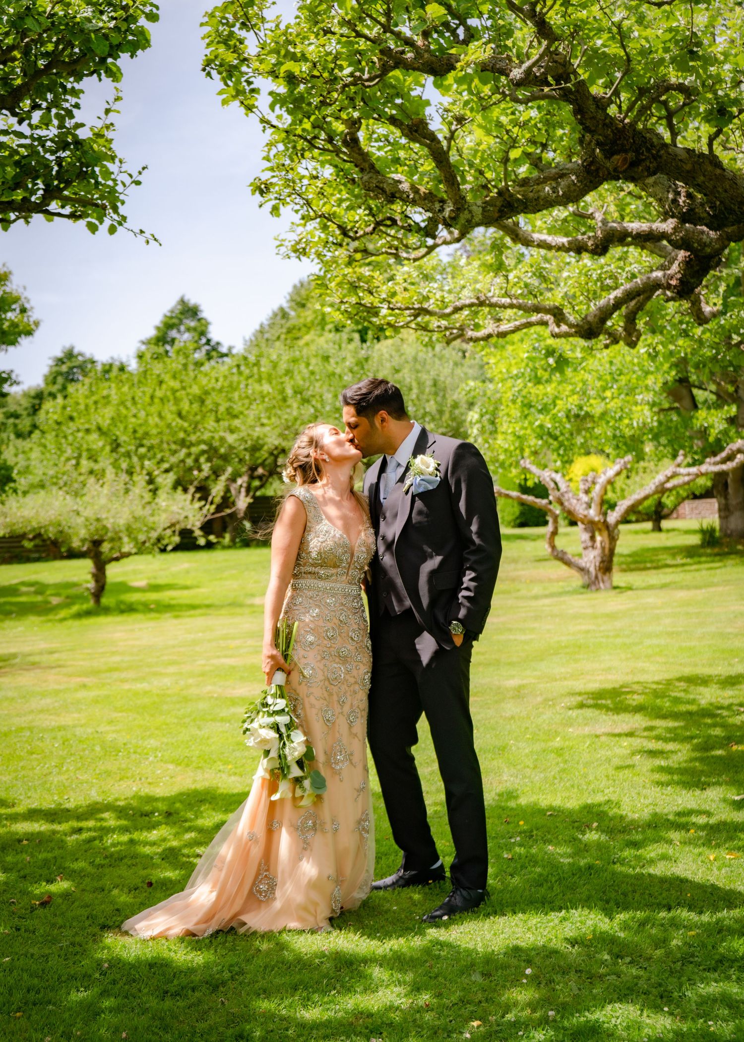 Bride in gold sequin gown and groom in black suit sharing a romantic kiss under a large oak tree.