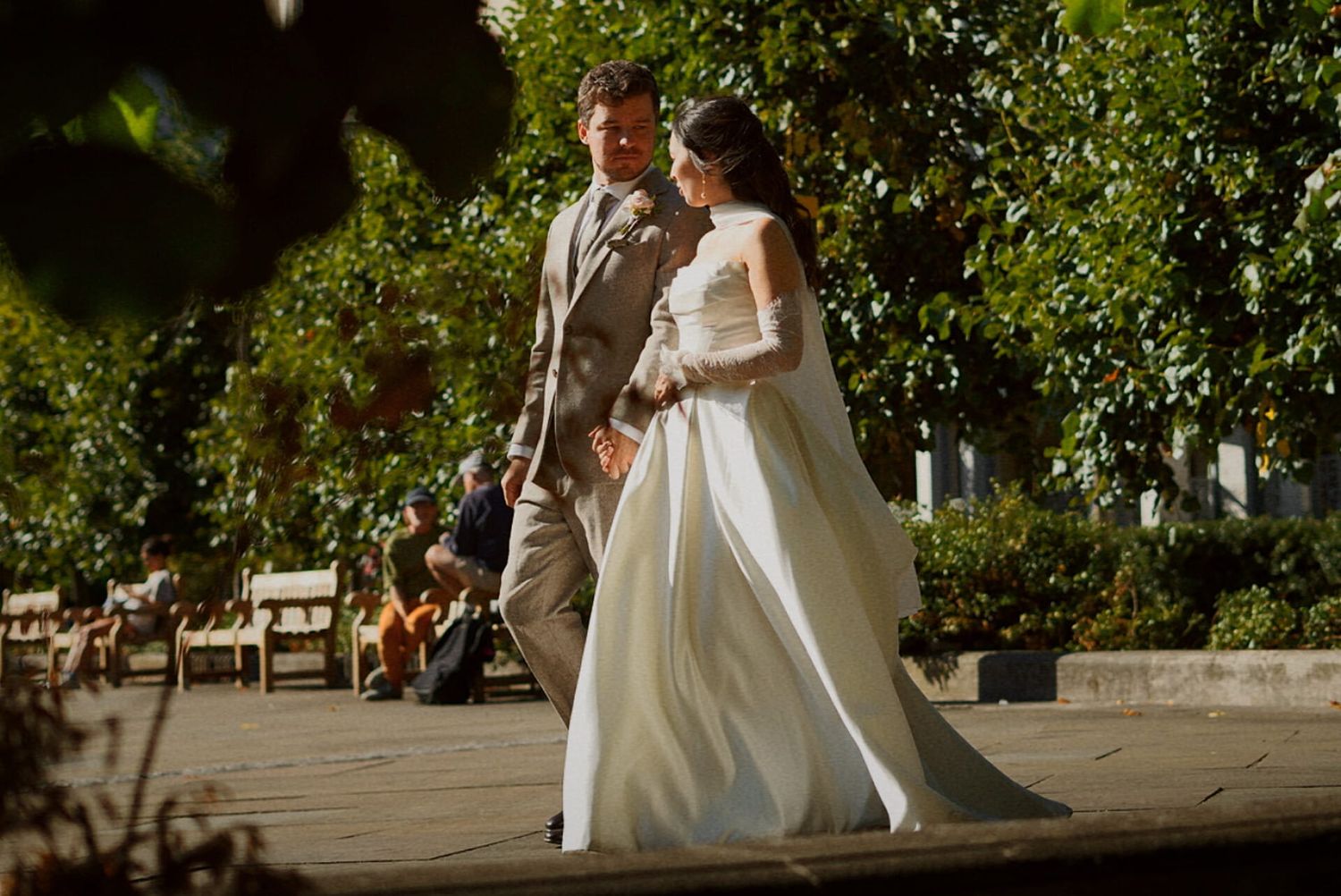 Bride in white gown and groom in grey suit walking together on a sunlit garden patio.