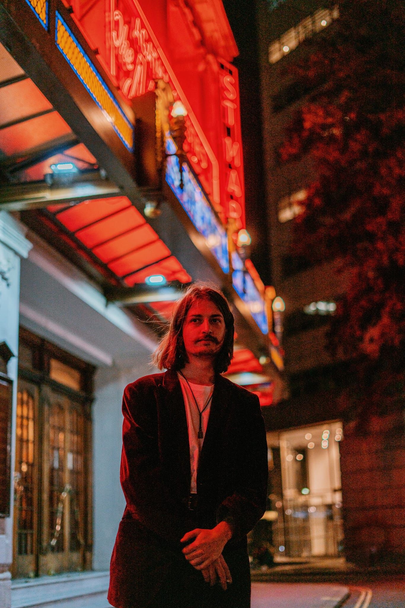 Man with long hair standing outside a lit-up building at night.
