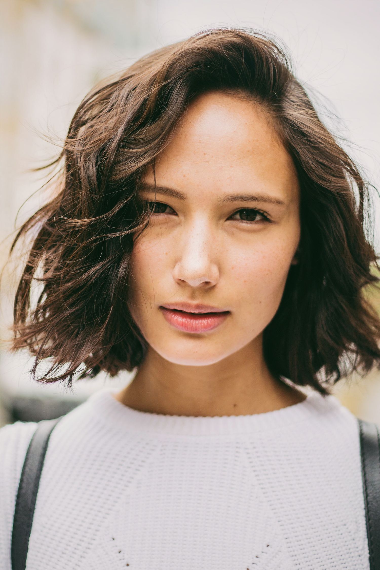 Young woman with short wavy brown hair wearing a white knit sweater outdoors in natural light.