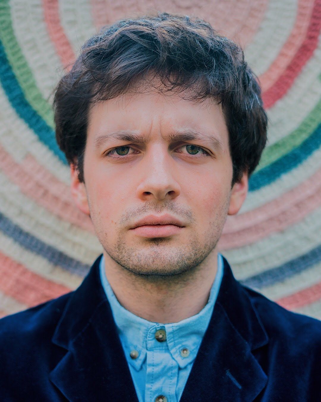 Dark-haired man in navy blazer and light blue shirt posing before colorful circular backdrop.