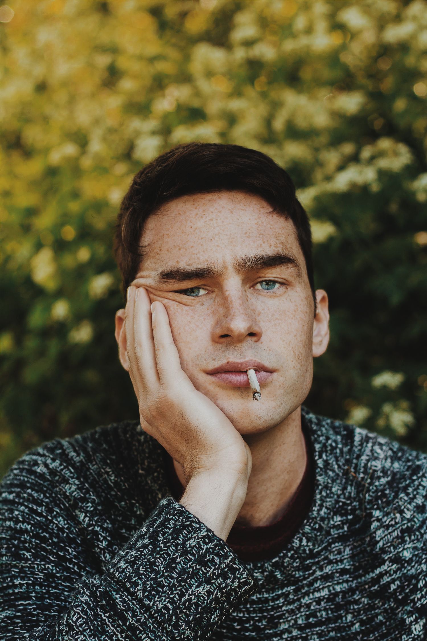 A freckled young man with blue eyes holds a cigarette while resting his cheek on his hand outdoors.