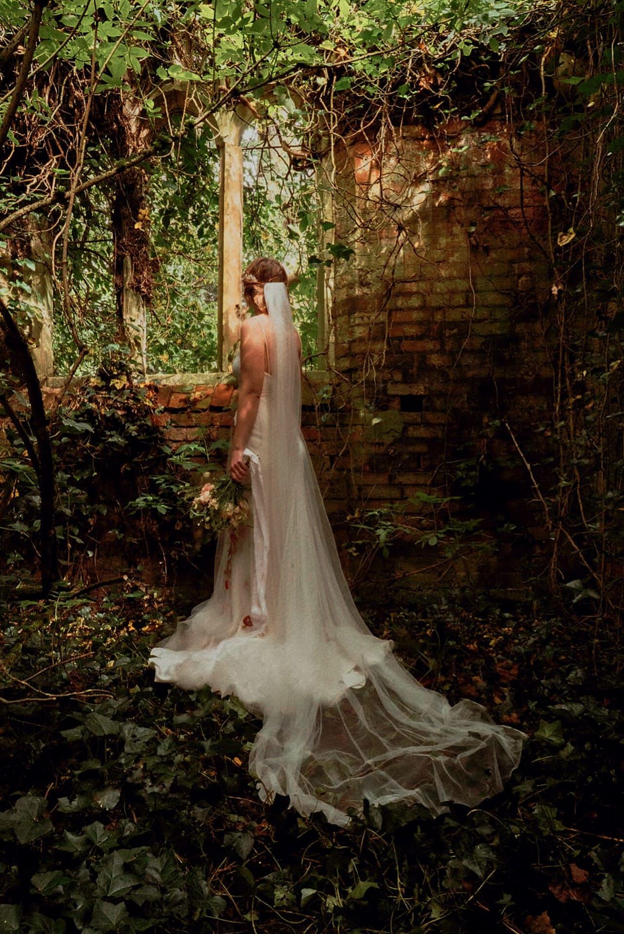 Bride in white wedding gown and veil standing in an ivy-covered ruined brick wall surrounded by lush greenery.