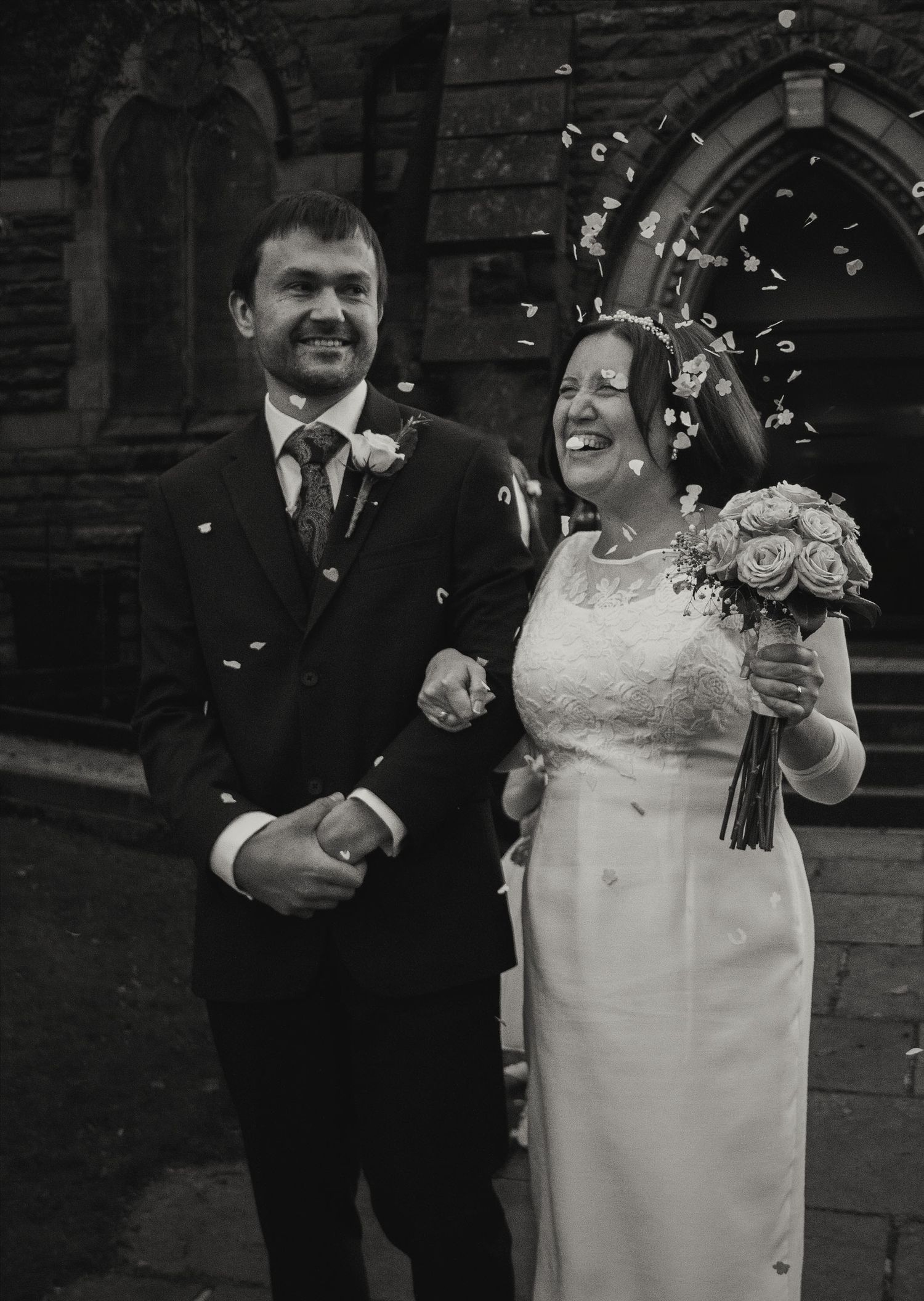 Smiling newlyweds in black and white photo with confetti outside historic stone church.