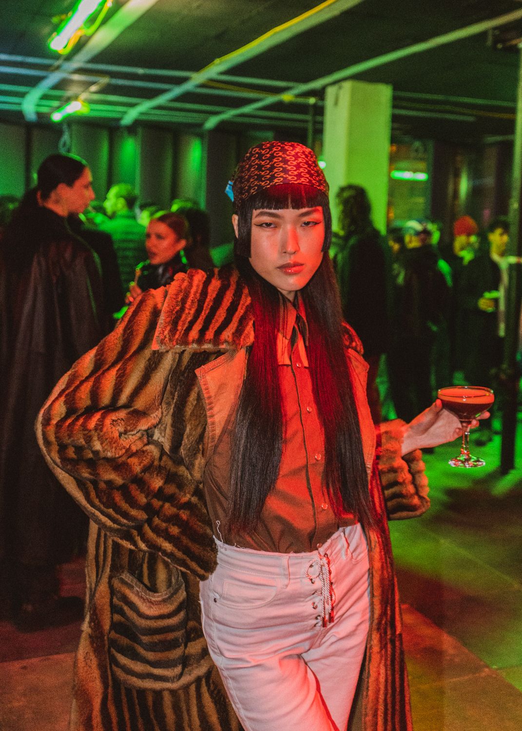 A stylish woman in a fur coat and red hat holds a cocktail at a neon-lit party.