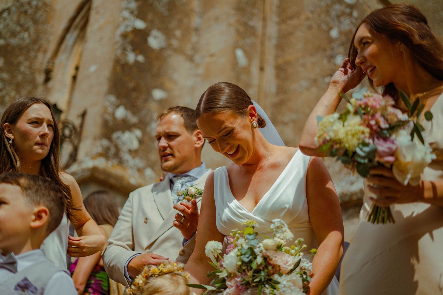 Emotional bride in white gown laughing and wiping tears while holding bouquet during outdoor wedding ceremony.