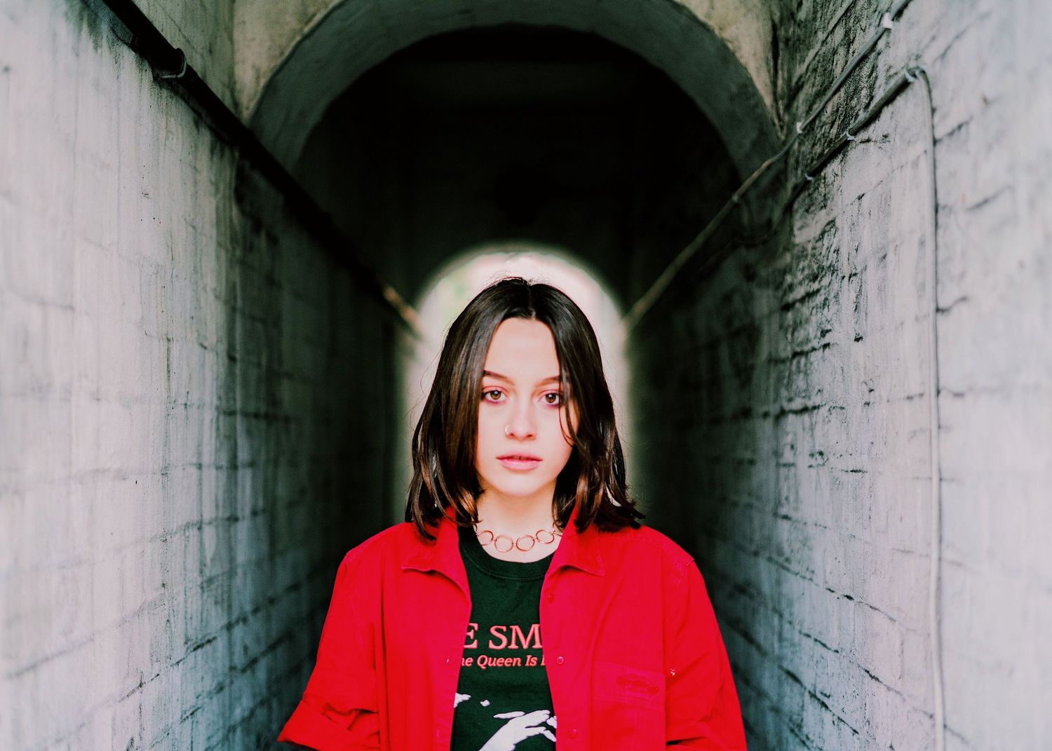 A young woman in a red jacket stands alone in a narrow concrete tunnel passageway.