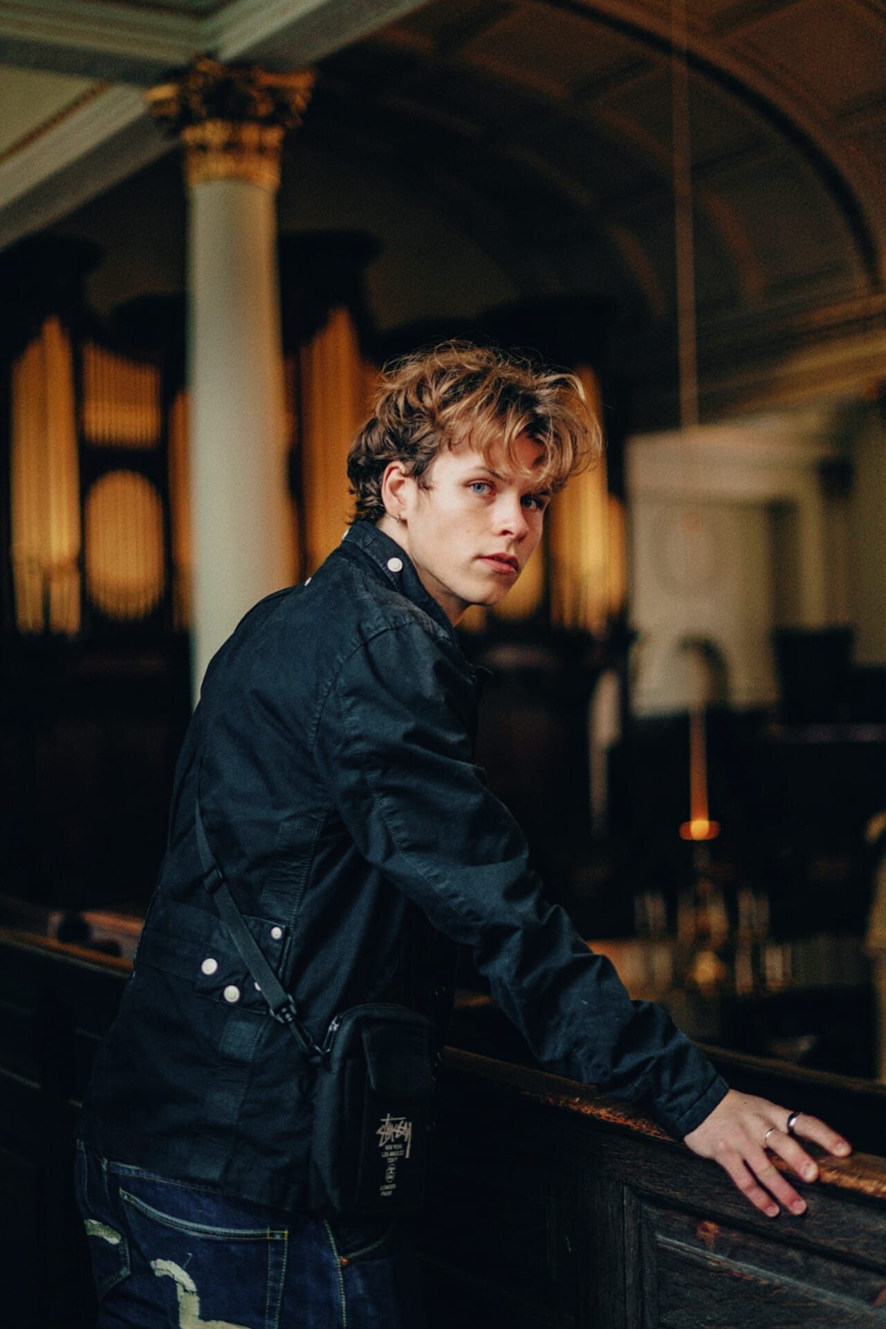 Young man with auburn hair in dark wax jacket leans on wooden pew in church.