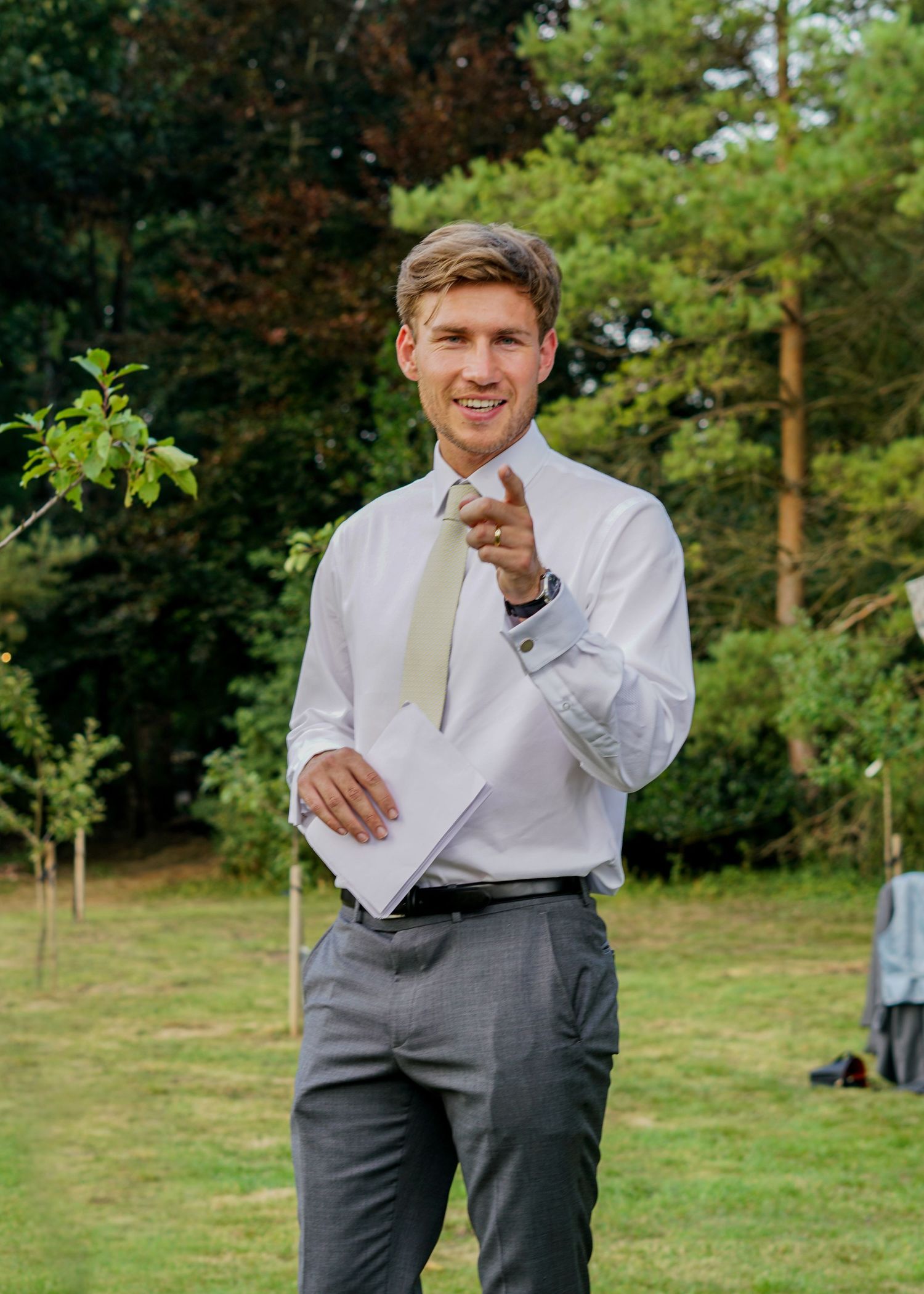 Smiling man in white shirt and gold tie pointing at camera during outdoor wedding.