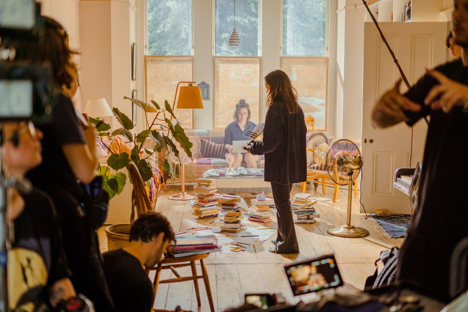 A film crew shoots a scene in a bright living room with a woman standing center frame.