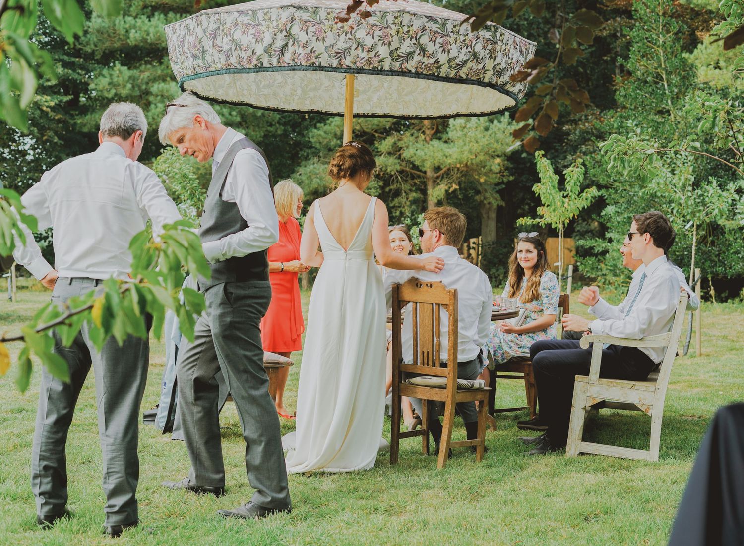 Wedding guests gathered outdoors under large umbrella at rustic garden reception table.