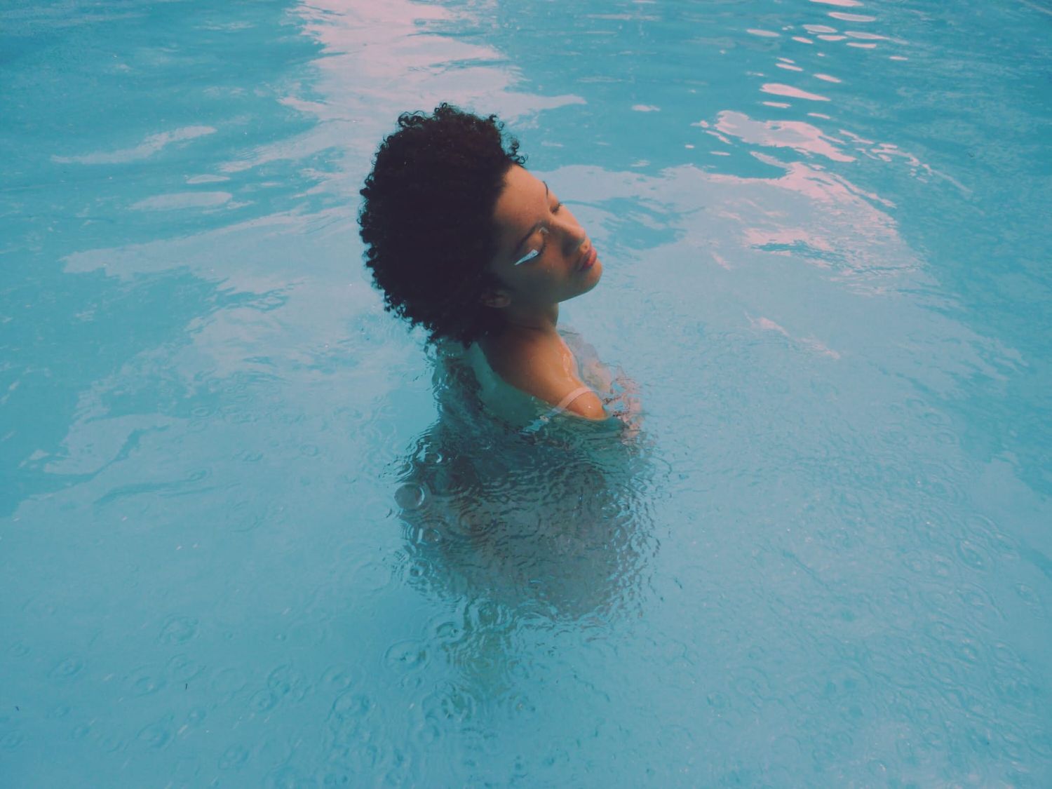 Person with natural curly hair relaxing in a bright blue swimming pool, head tilted back.