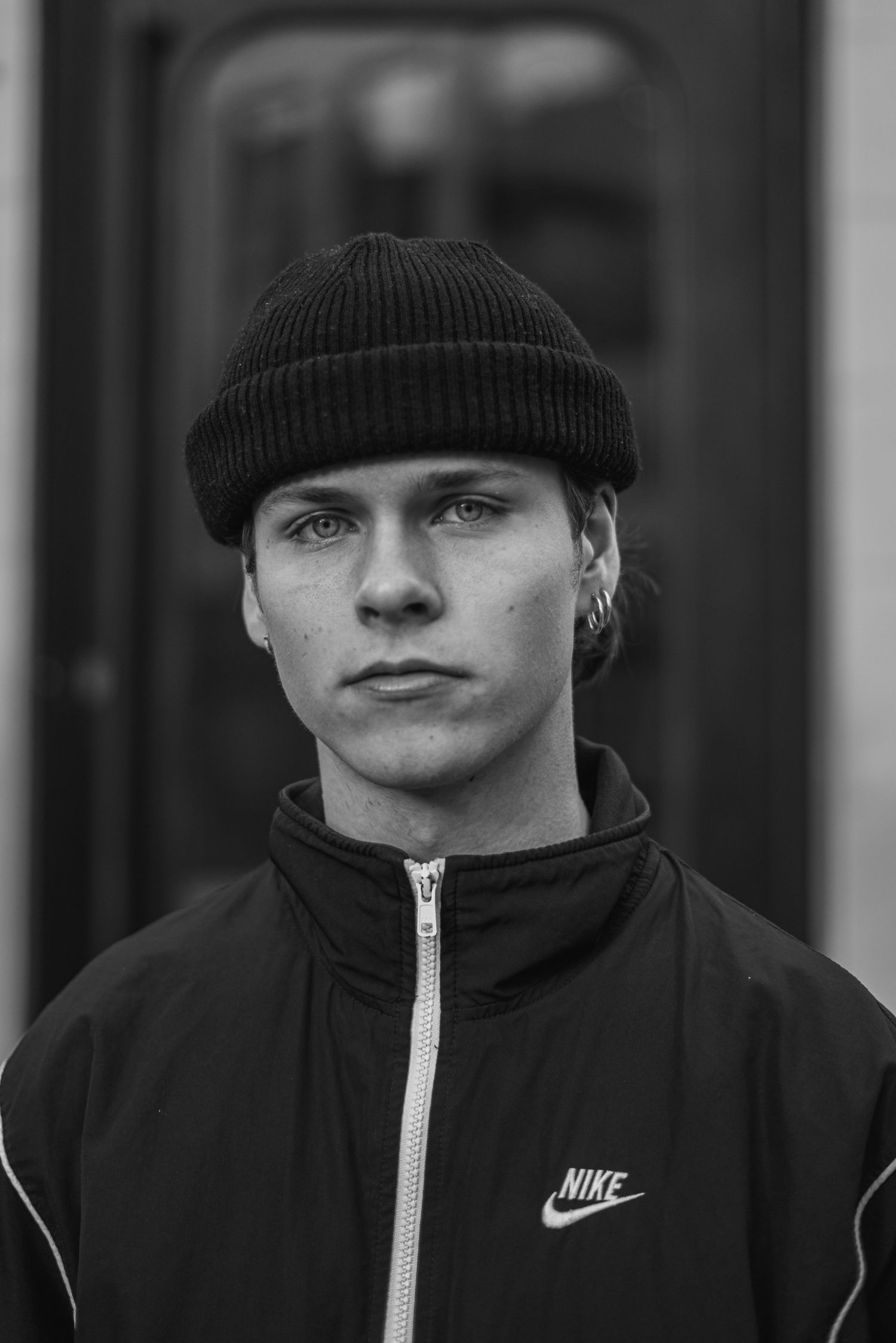 Young man in a black Nike jacket and beanie hat posing for a black and white portrait.