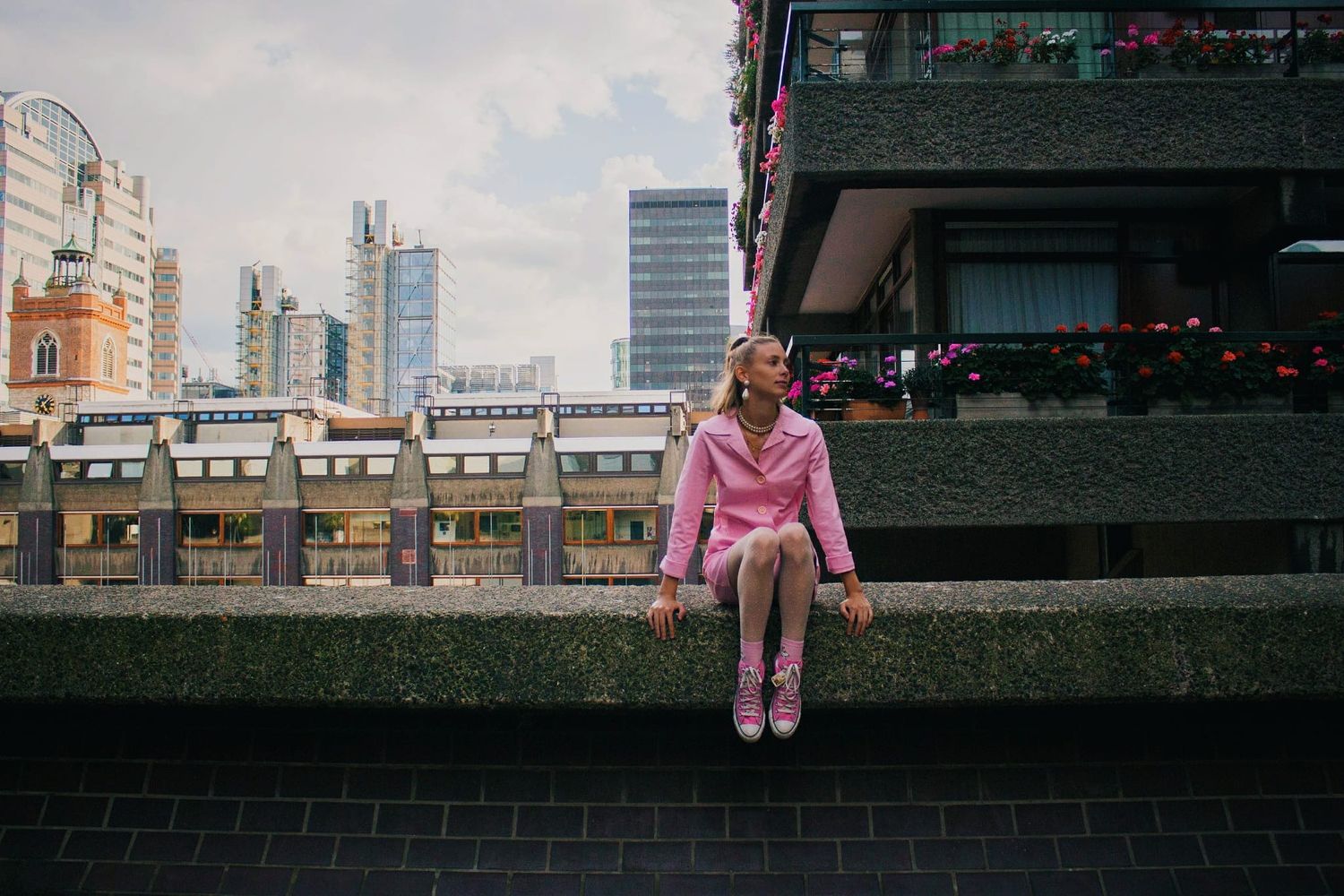 Woman in pink outfit sitting on urban wall with city skyline behind her.