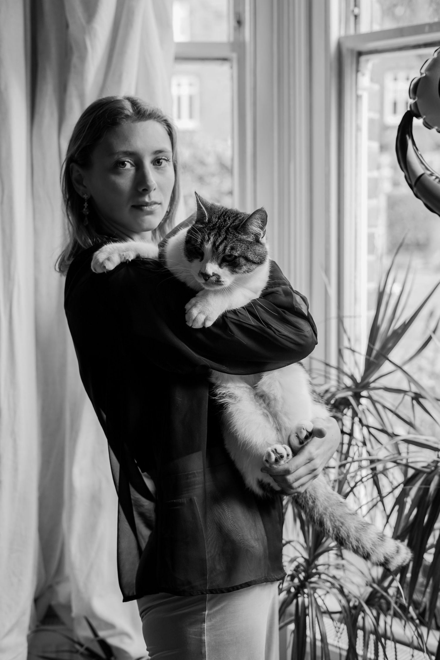 Black and white portrait of blonde woman holding large fluffy cat near window with plants.
