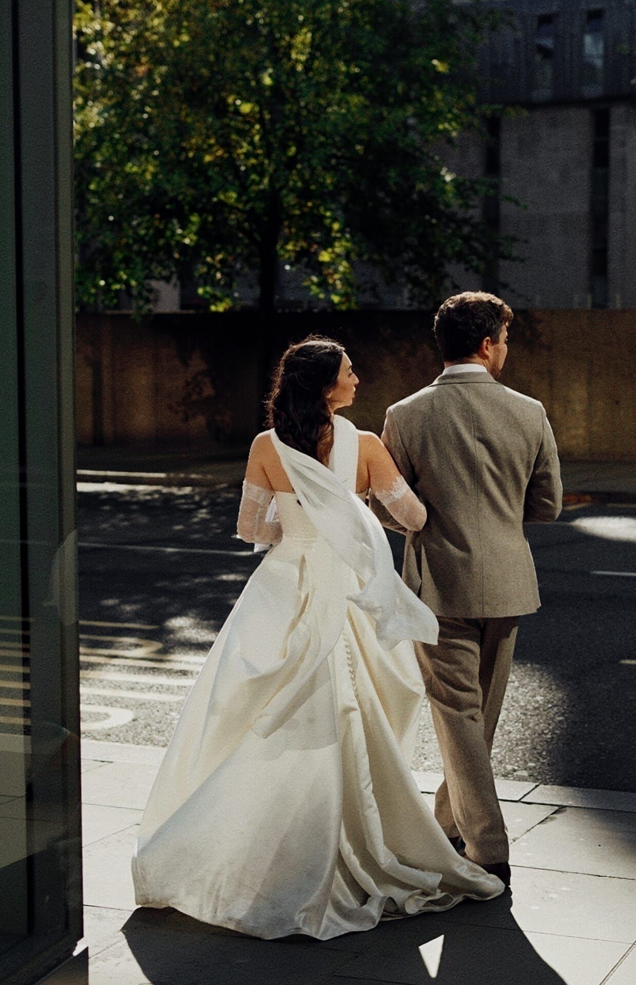 Bride in elegant white ballgown and groom in tan suit walking toward glass doors in sunlight.