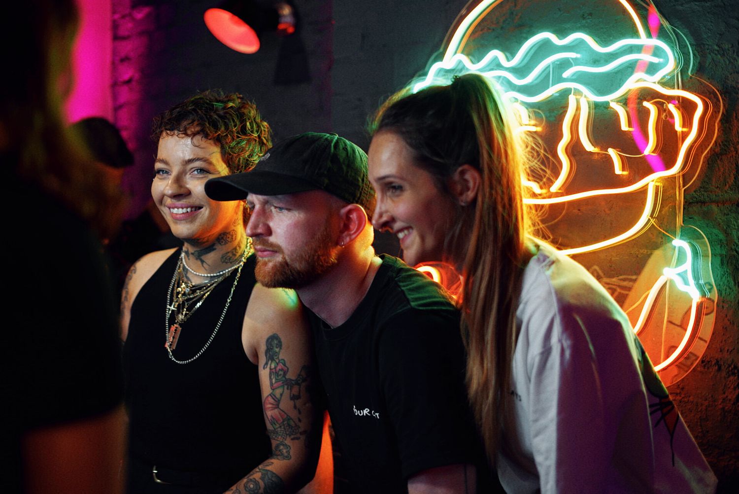 Three friends laugh together beneath a glowing neon sign at an indoor venue.
